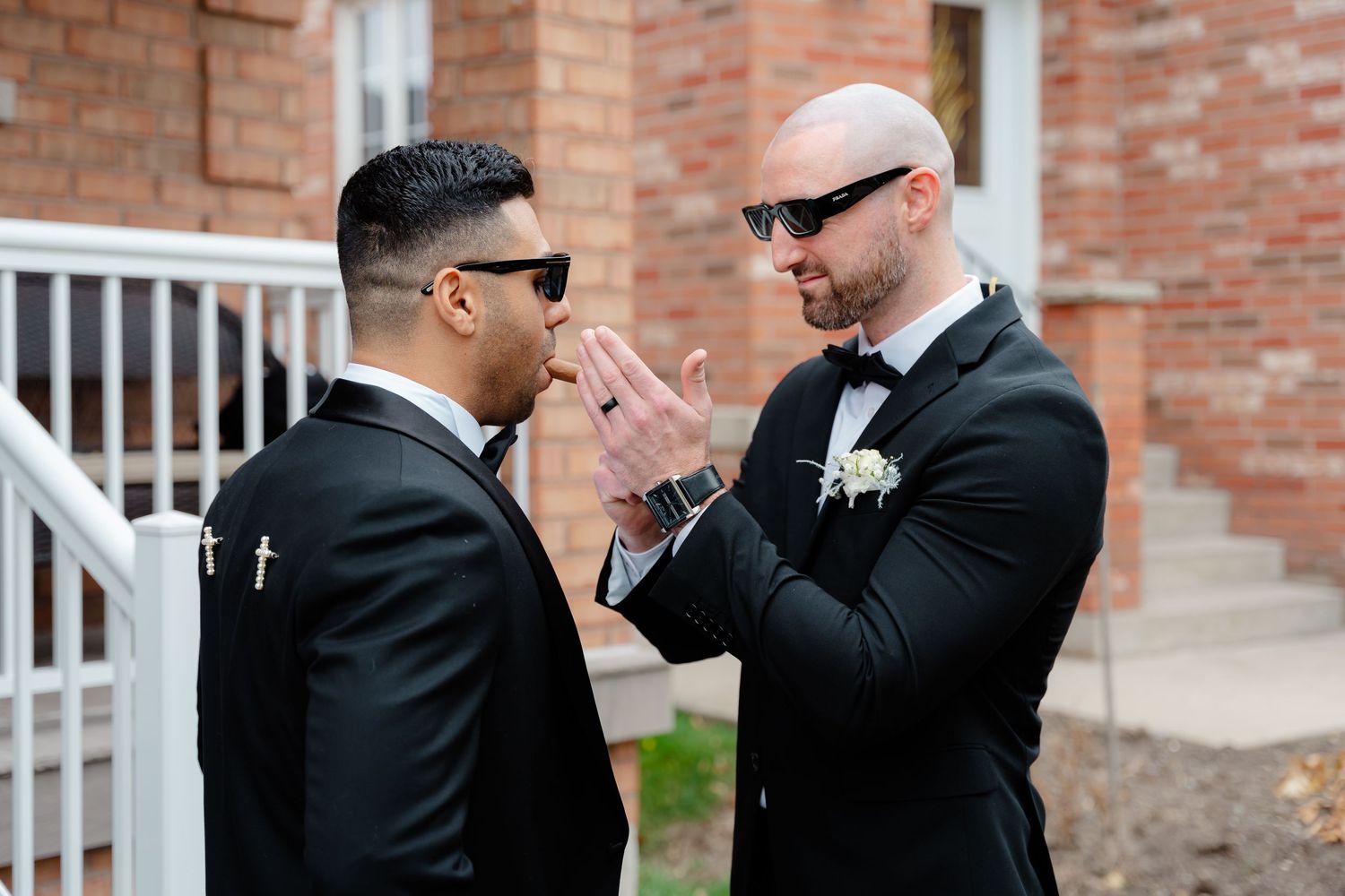 Two groomsmen in black tuxedos and sunglasses sharing an animated moment outside.
