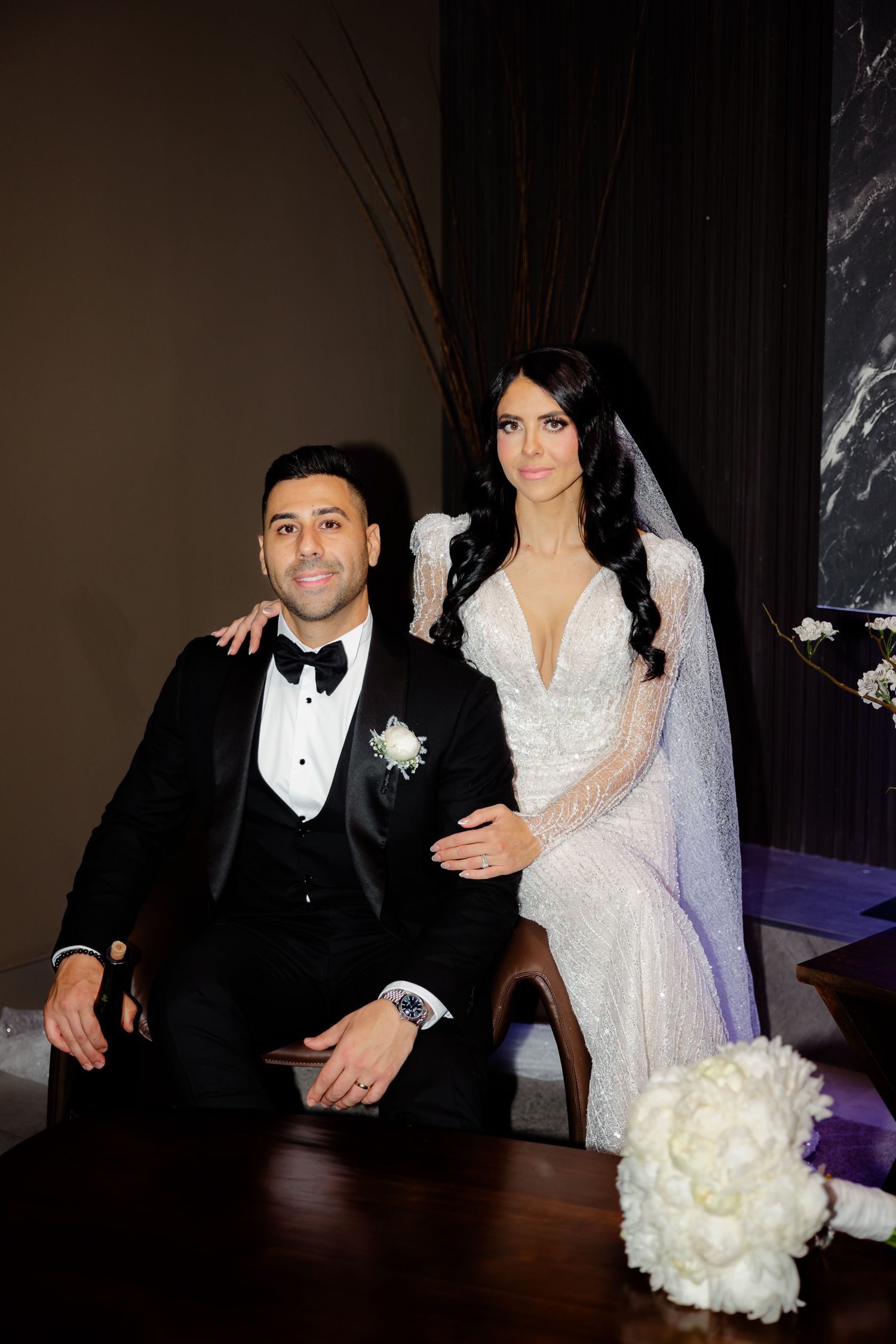 Bride in white lace dress stands behind seated groom in formal tuxedo during wedding photoshoot.