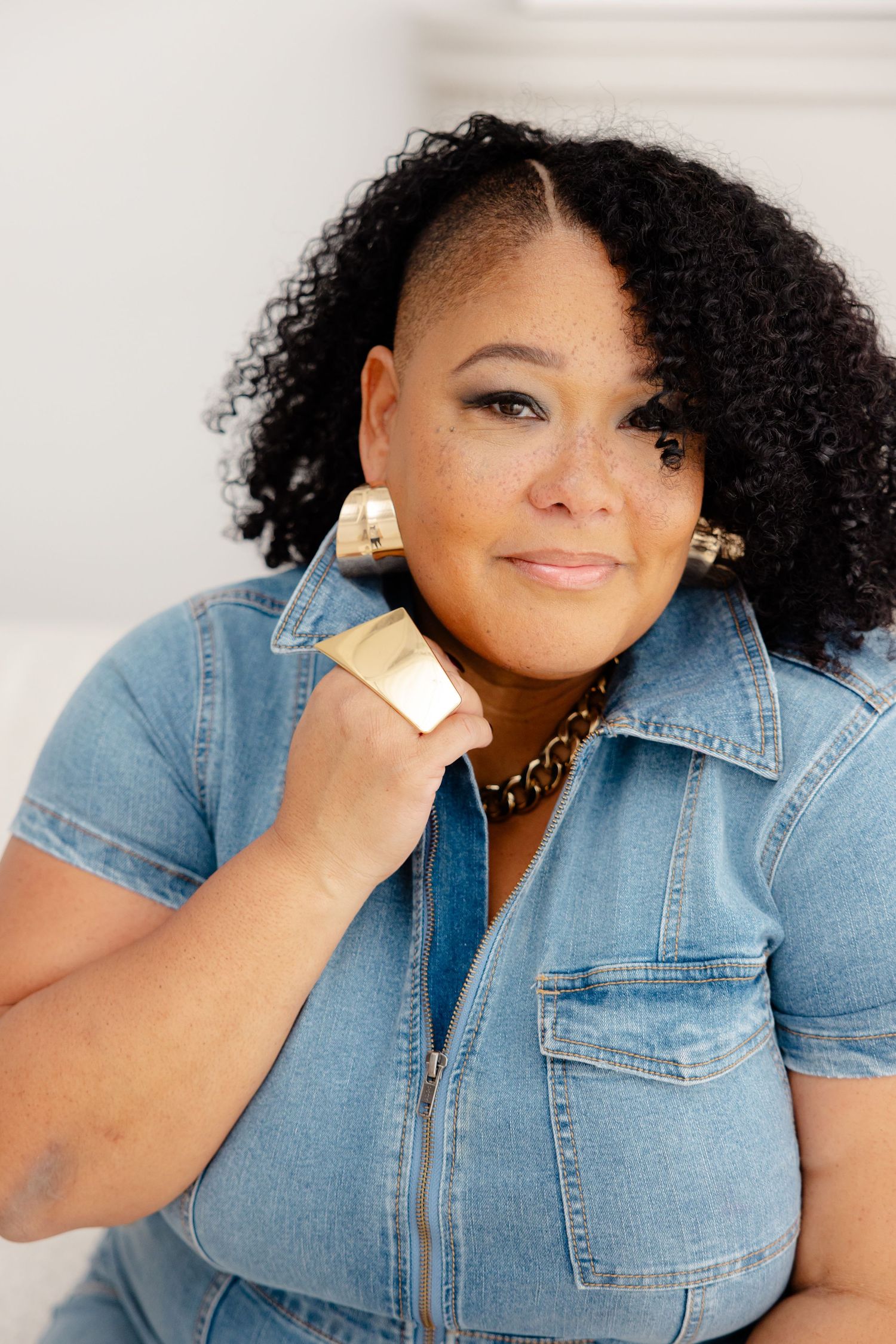 Smiling woman with natural curly hair wearing denim shirt and statement jewelry poses against light background.