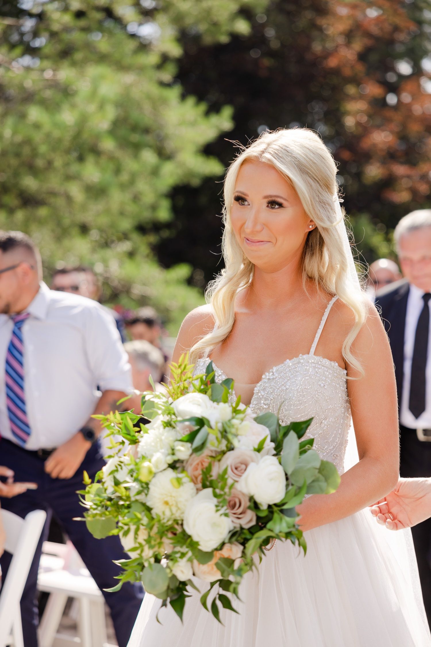 A bride in a delicate spaghetti strap gown holds a white and green bouquet during an outdoor ceremony.