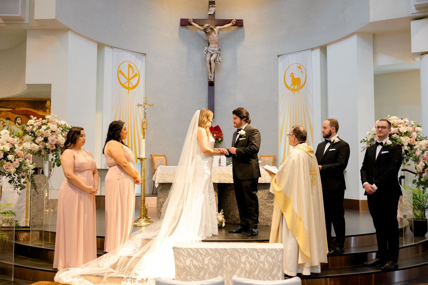 Wedding ceremony in progress with priest officiating and bridesmaids in blush dresses.