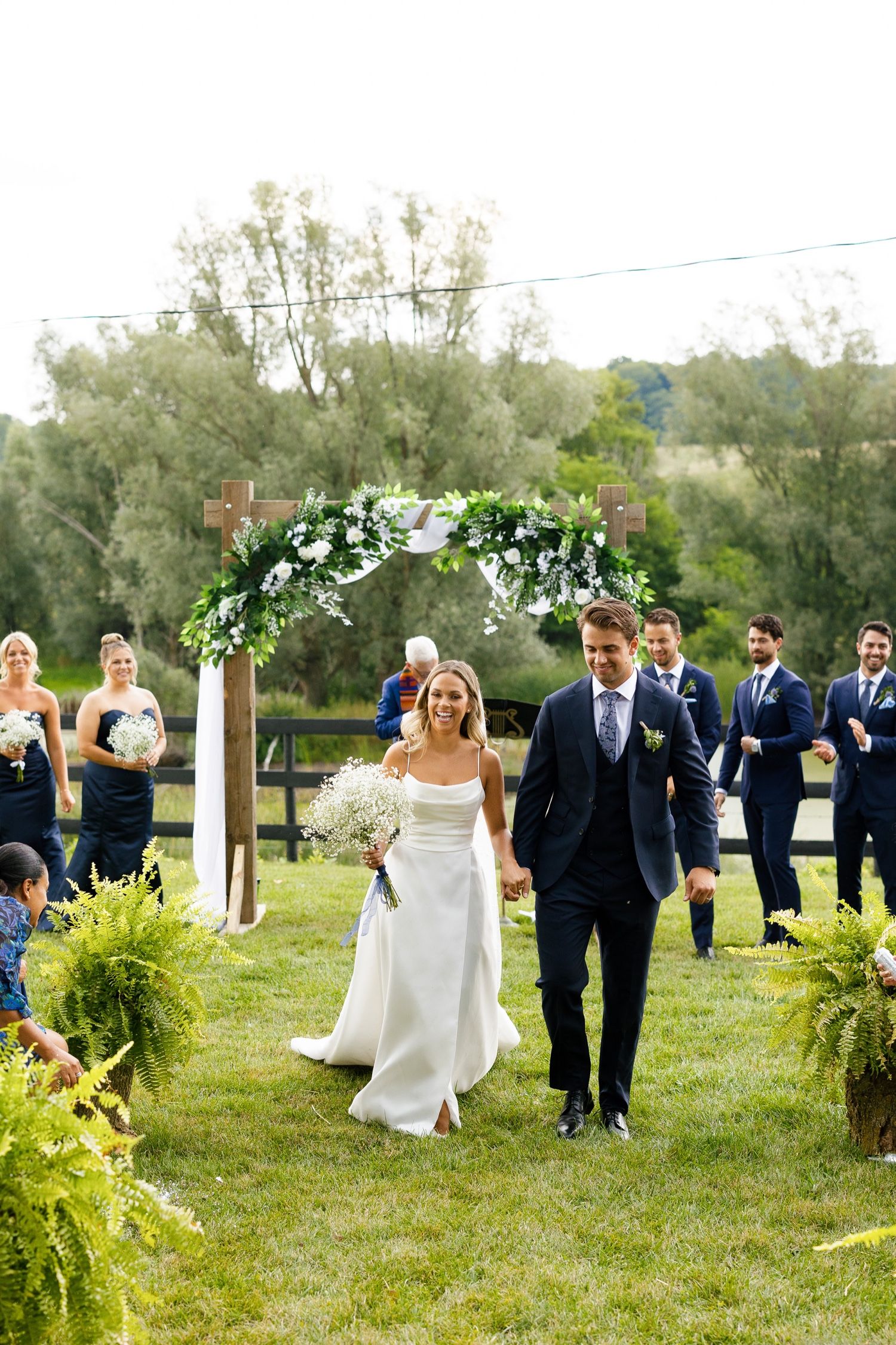 A couple walks through their outdoor wedding venue decorated with lush ferns and a floral archway.