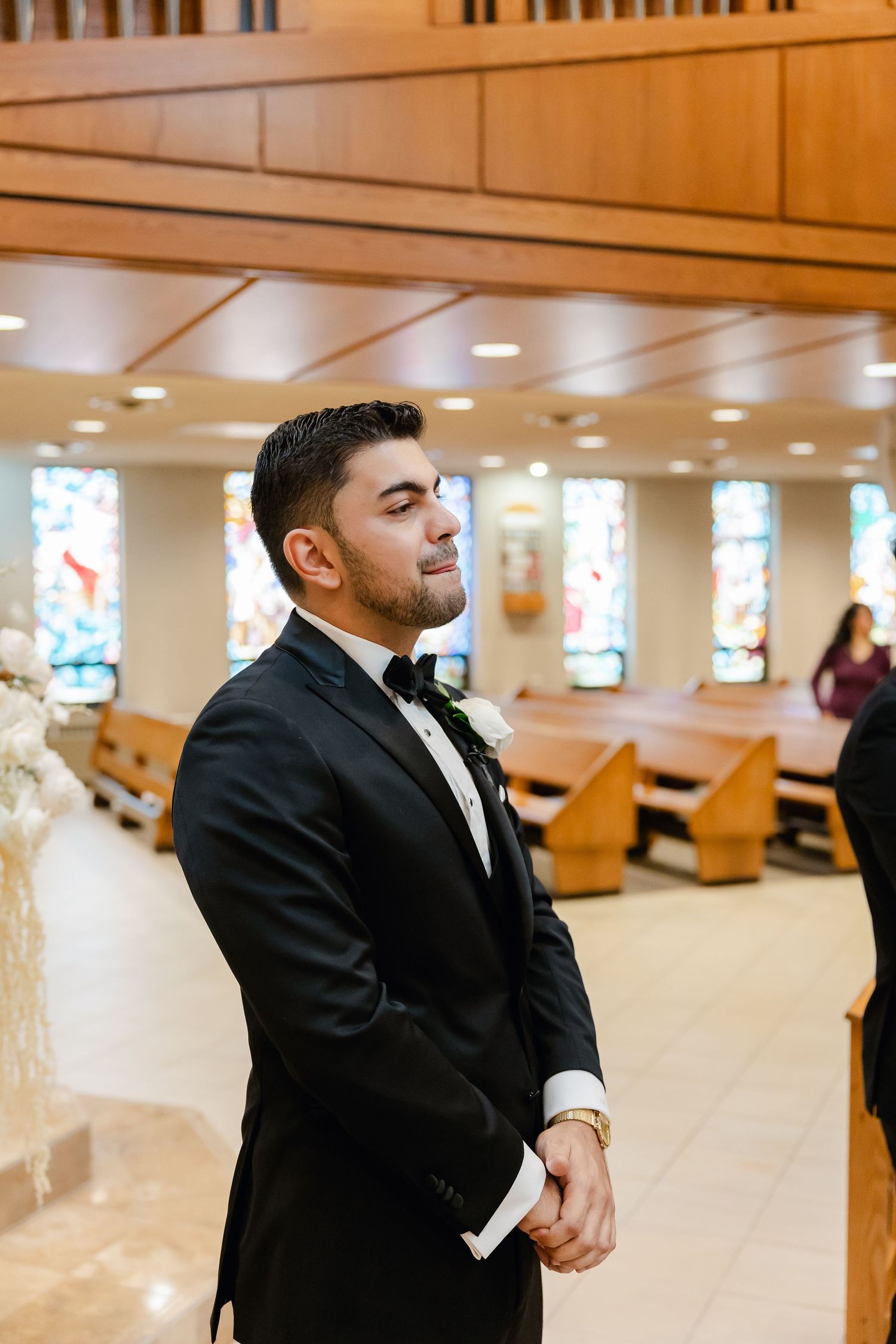 Groom in classic black tuxedo and bowtie waits in church pews during wedding ceremony.