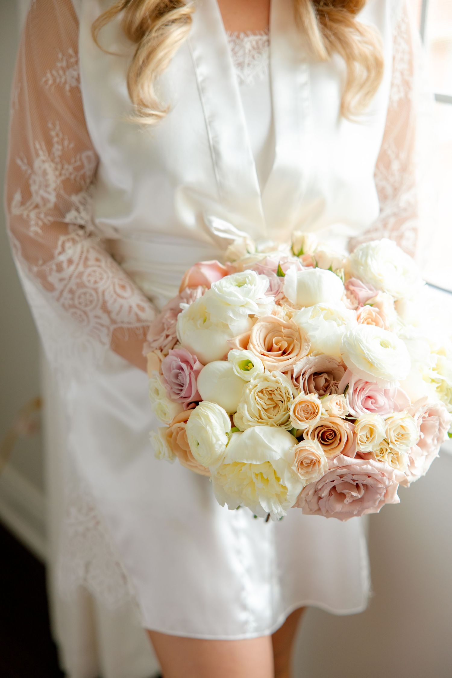 A romantic bridal bouquet with blush pink and white roses held against a white lace dress.