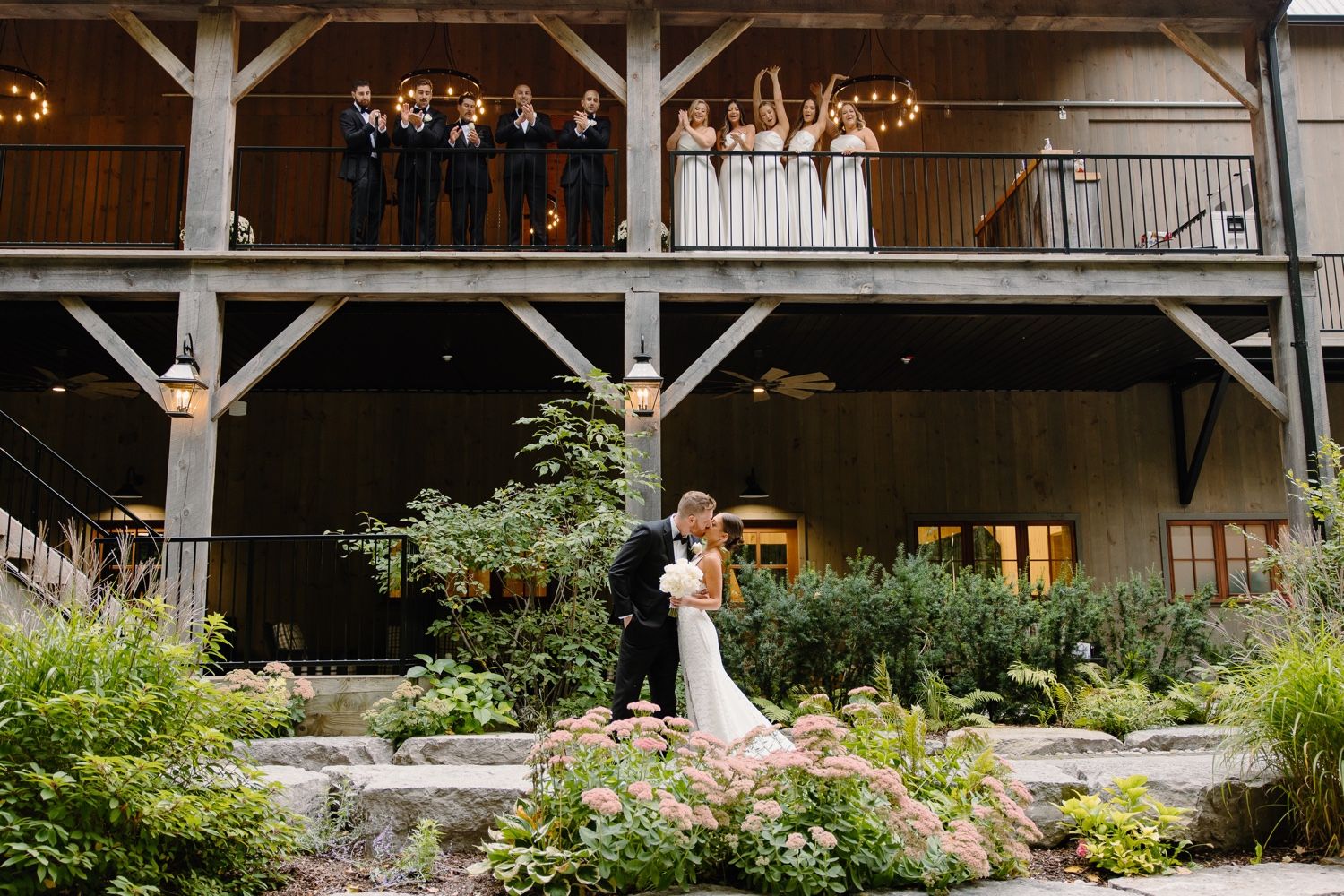 Elegant wedding photo with couple on garden path and wedding party on rustic venue balcony.