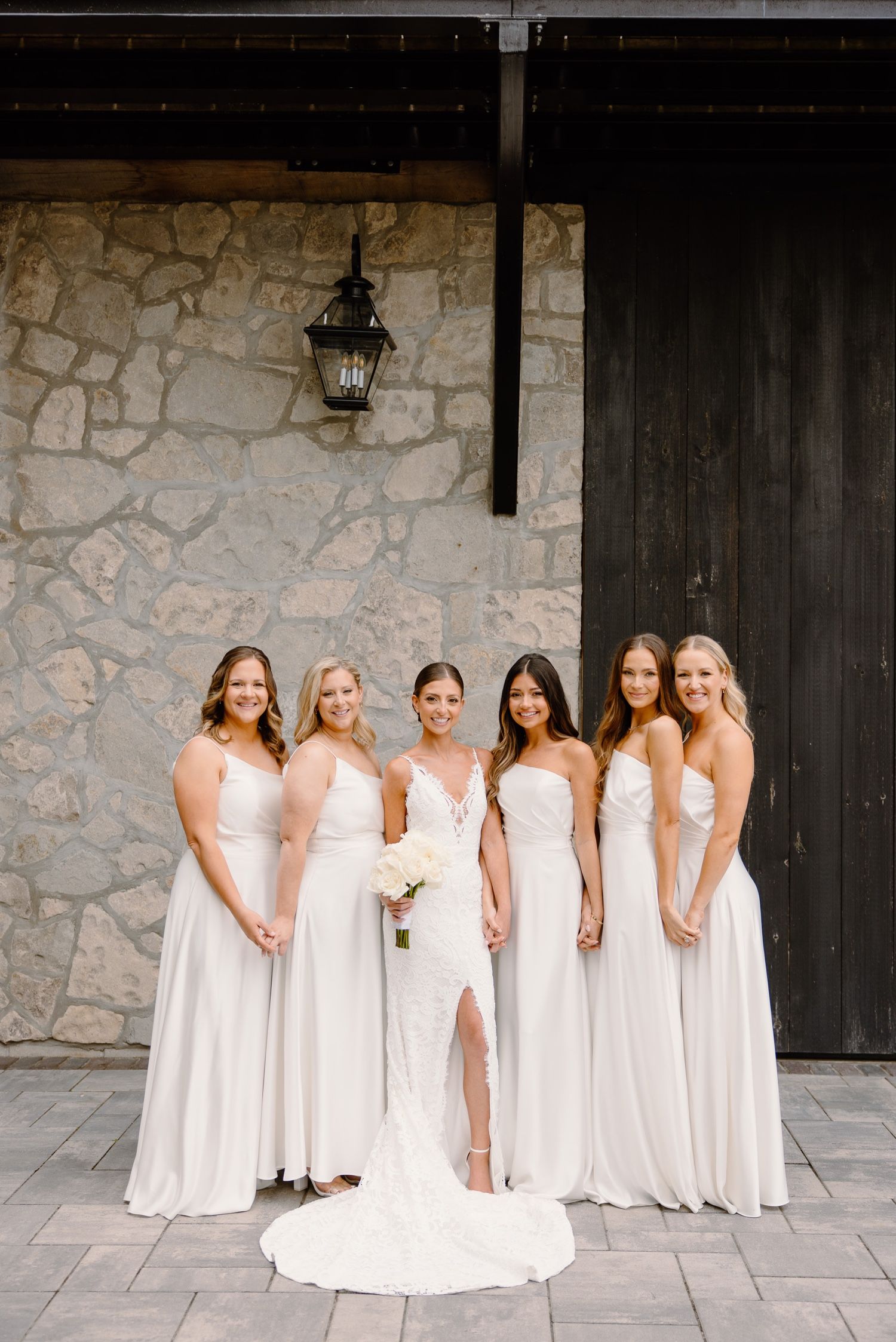 Wedding party in coordinating white gowns posed formally against stone wall with lantern.