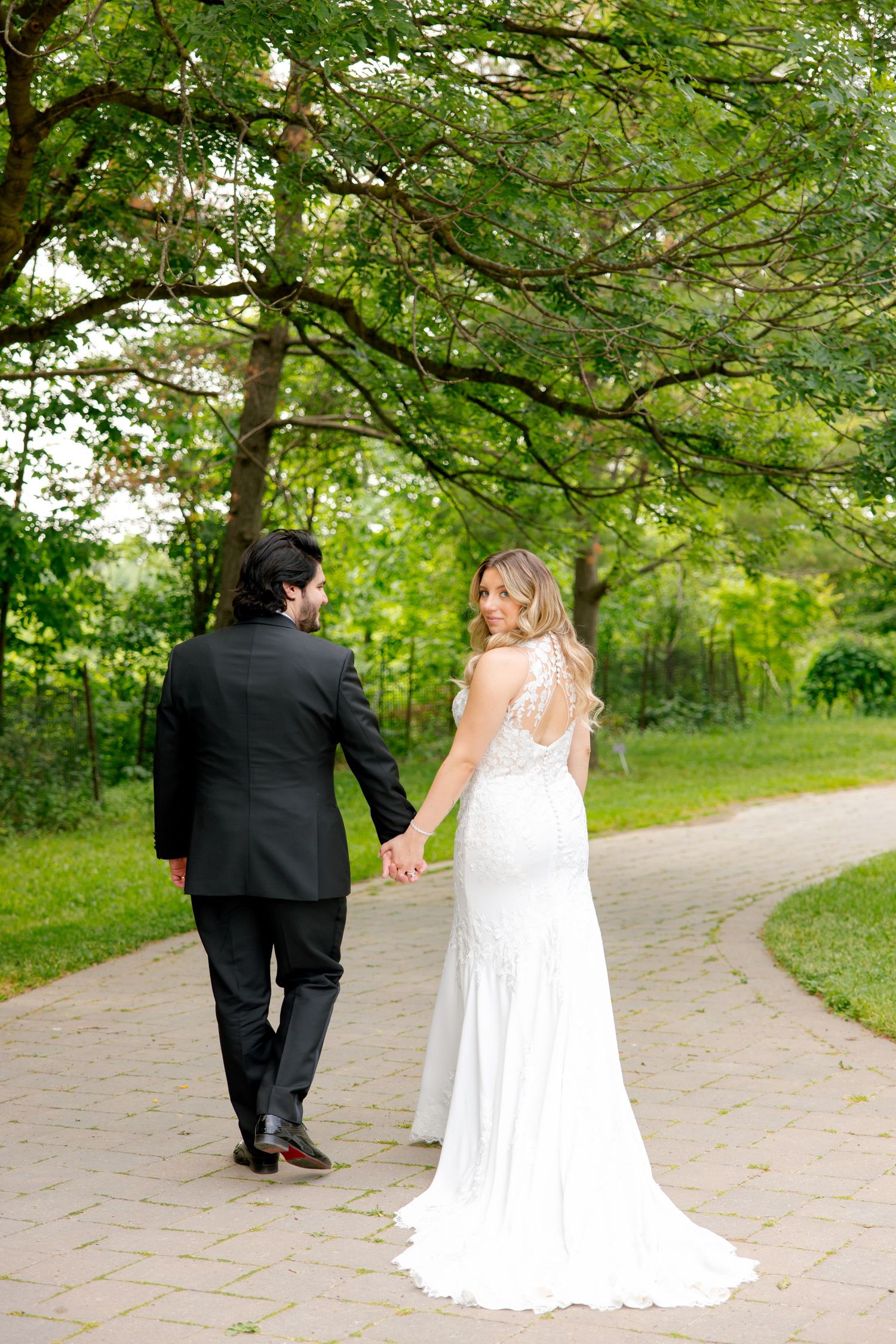 Newlyweds walking hand in hand down a garden path surrounded by lush green trees.