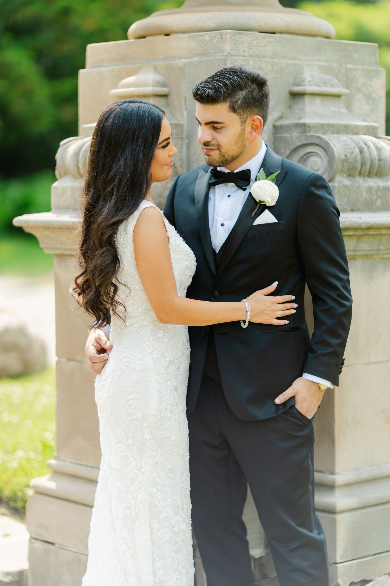 Elegant wedding couple poses against ornate stone pillar in verdant garden setting.