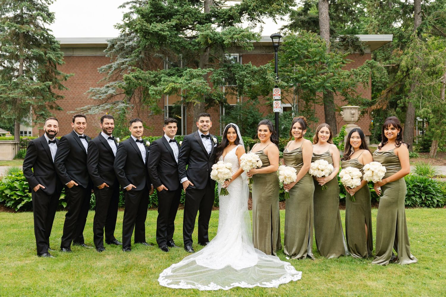 Formal wedding party portrait with bridesmaids in sage green and groomsmen in black suits on a lawn.