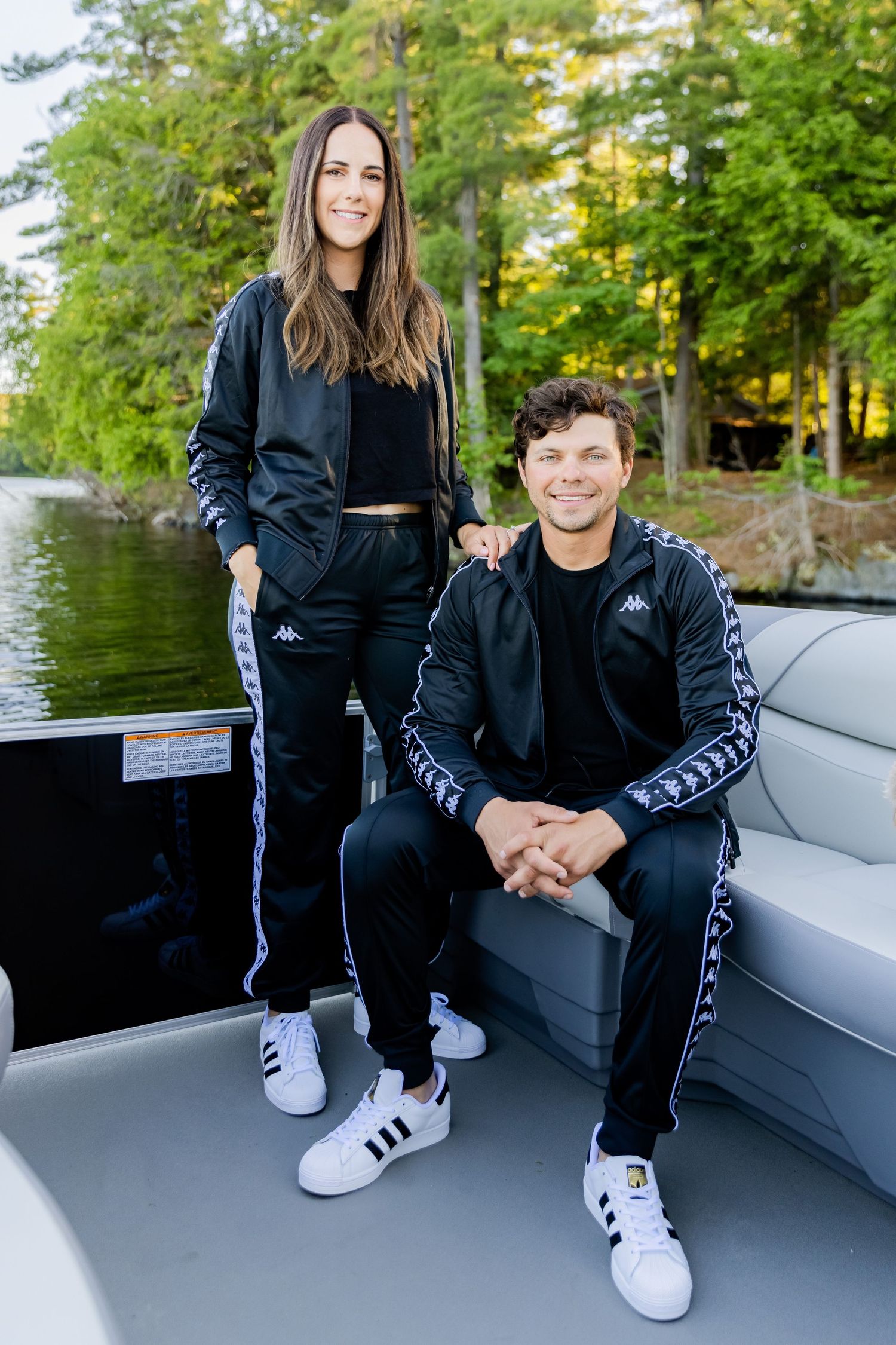 A couple wearing matching black and white Kappa athletic wear pose on a boat with trees in background.