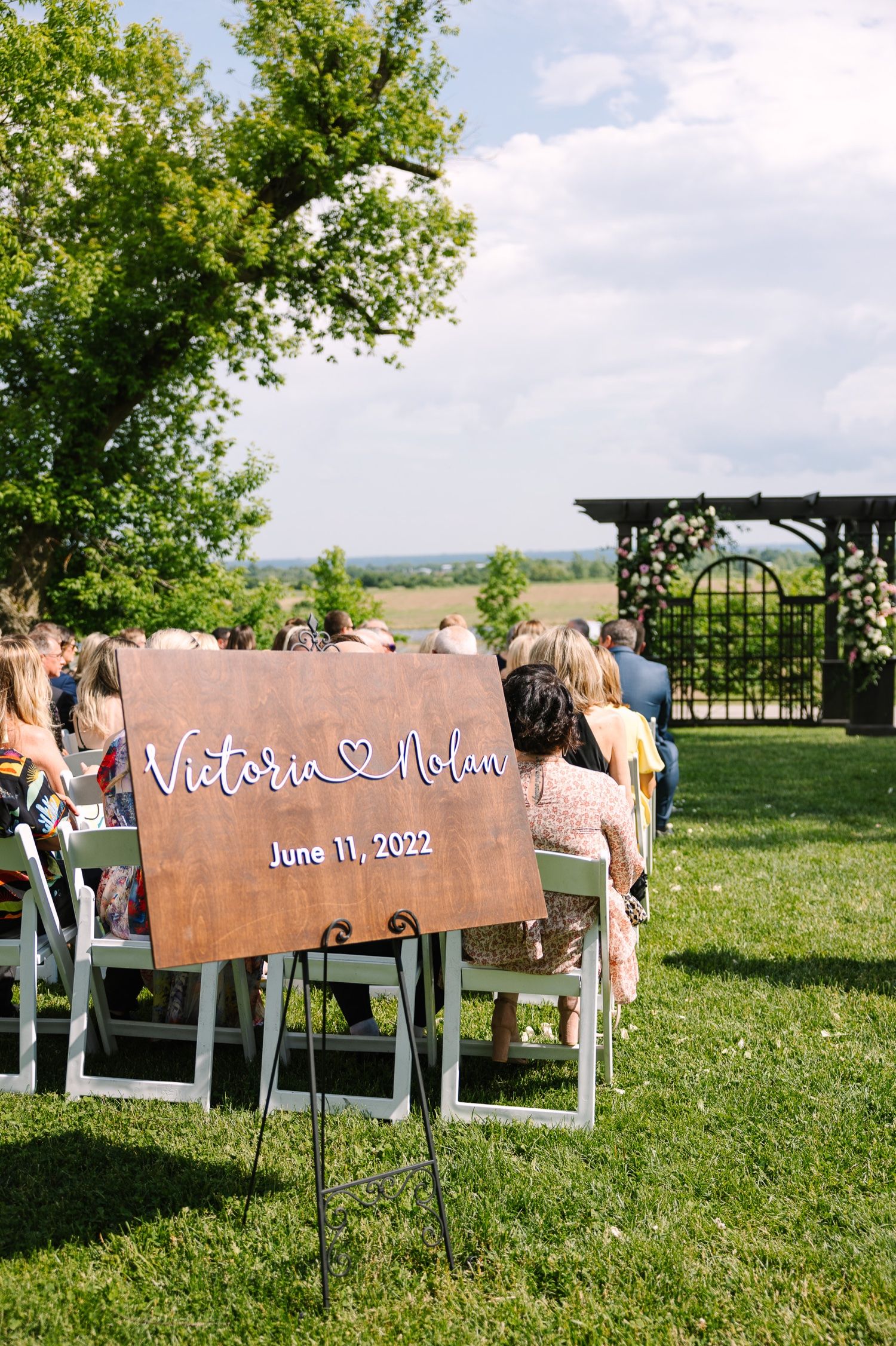 Rustic wooden wedding welcome sign displayed on lawn with ceremony seating in background.
