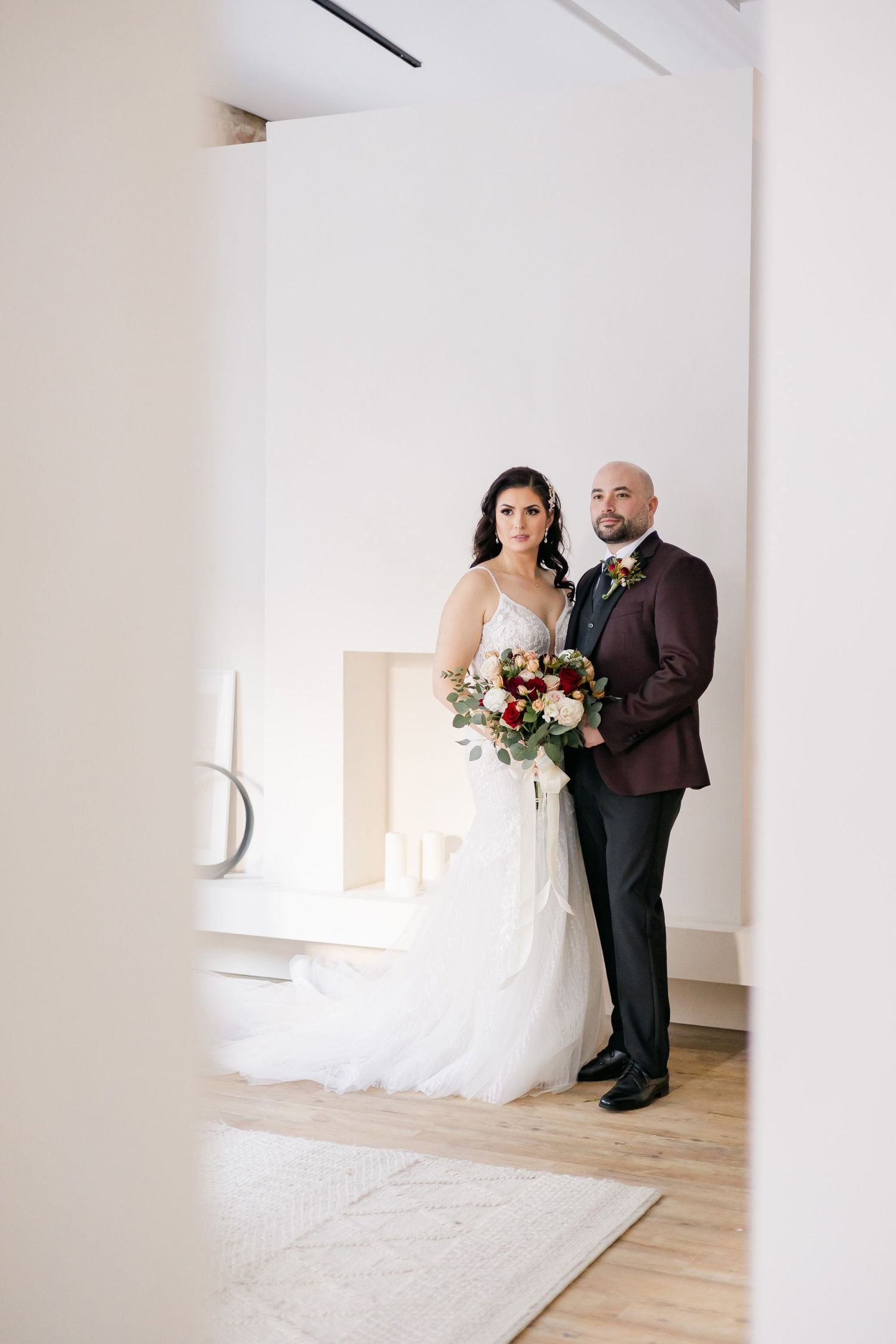 Elegant wedding portrait featuring burgundy suit and white gown with vibrant red and white bouquet.