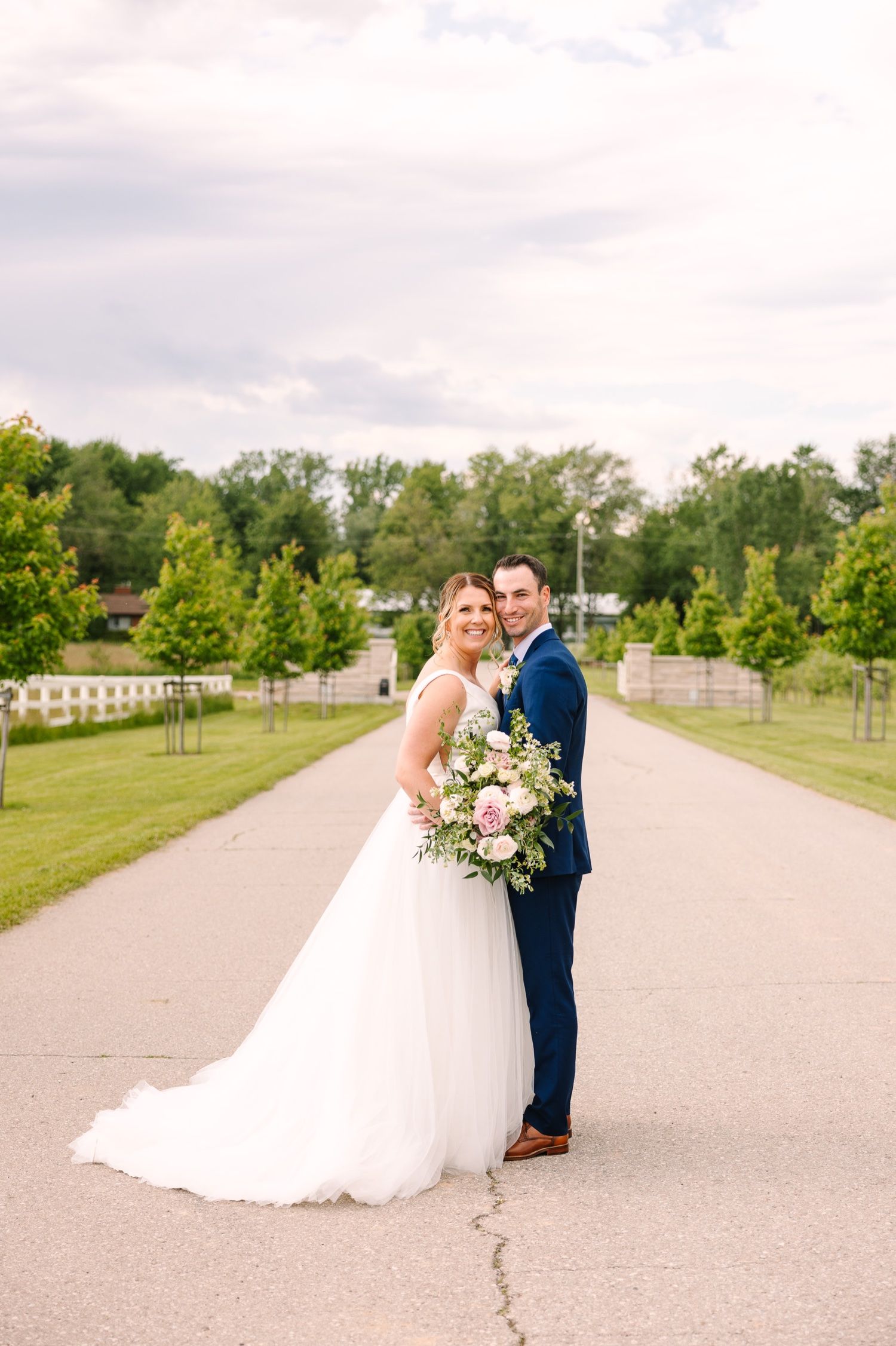 Wedding couple embraces on tree-lined path with beautiful summer lighting.
