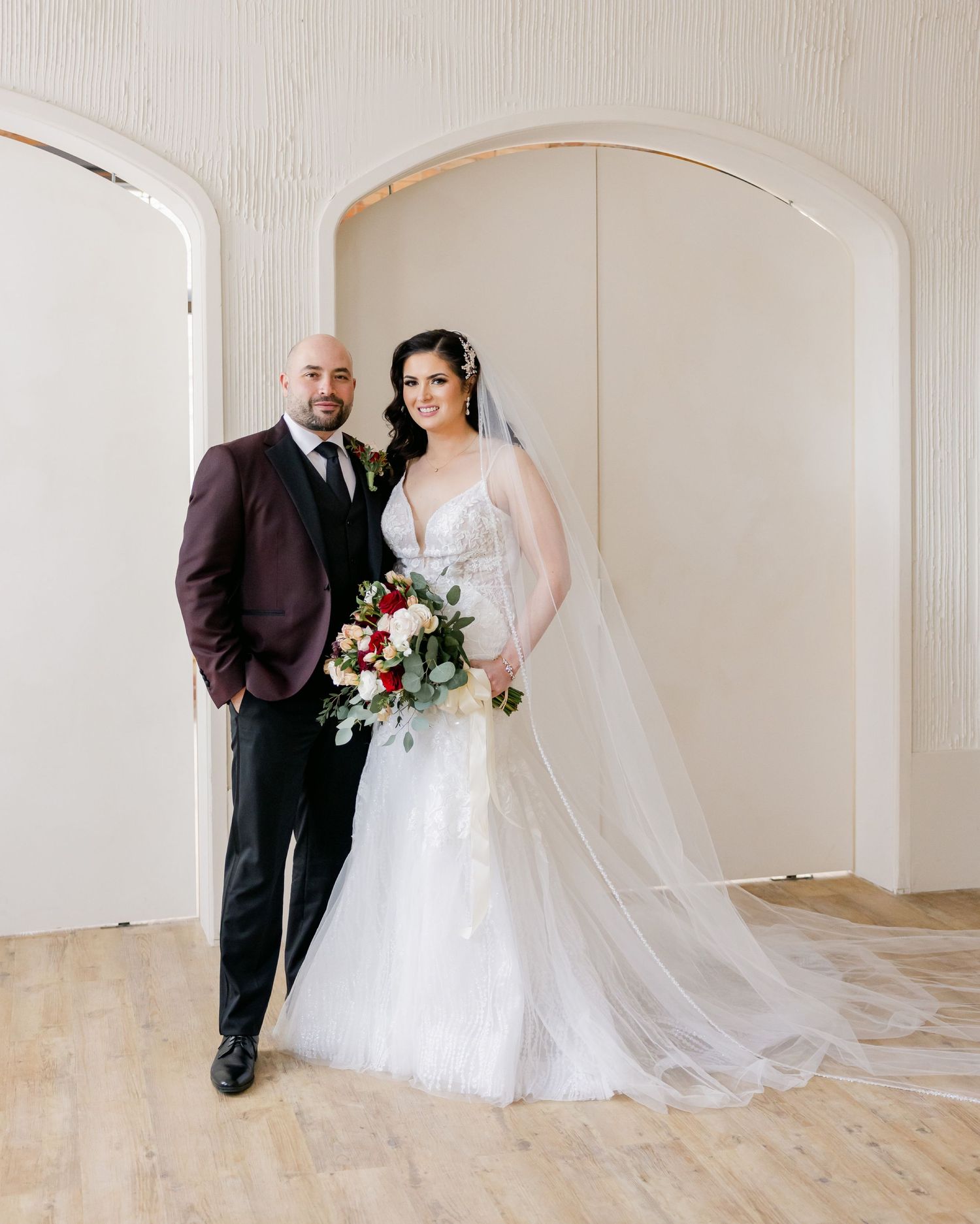 Classic wedding portrait featuring burgundy suit and white lace gown under arched doorway.