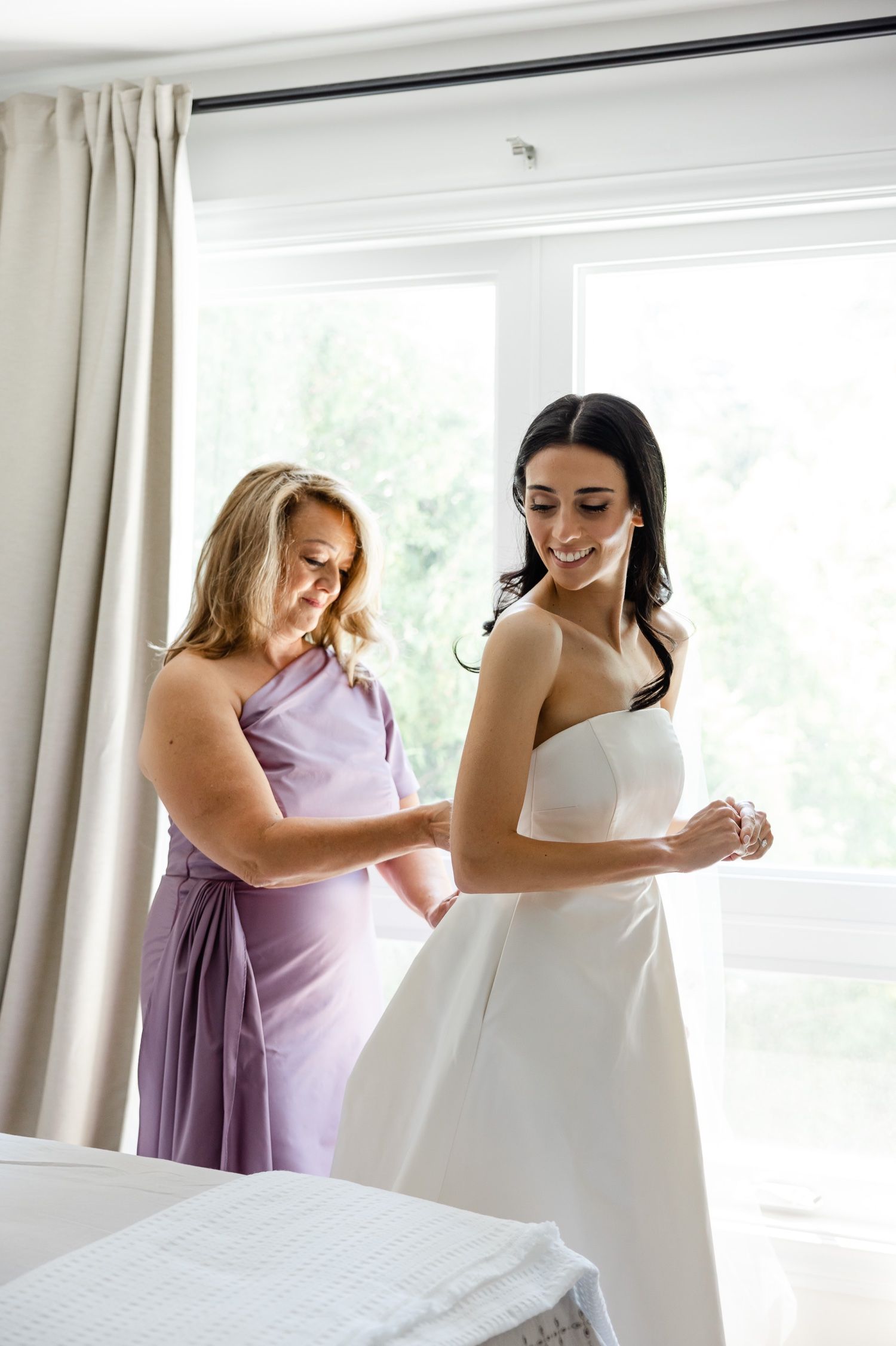 Final dress adjustments being made in sunlit bedroom on wedding morning.