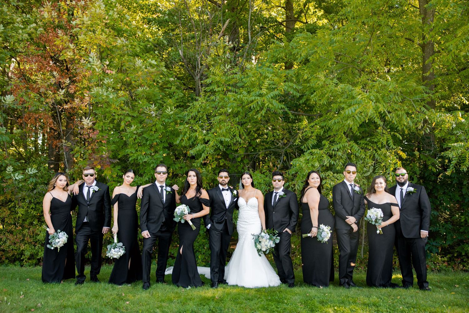 Wedding party in formal black attire poses together against a backdrop of autumn foliage.