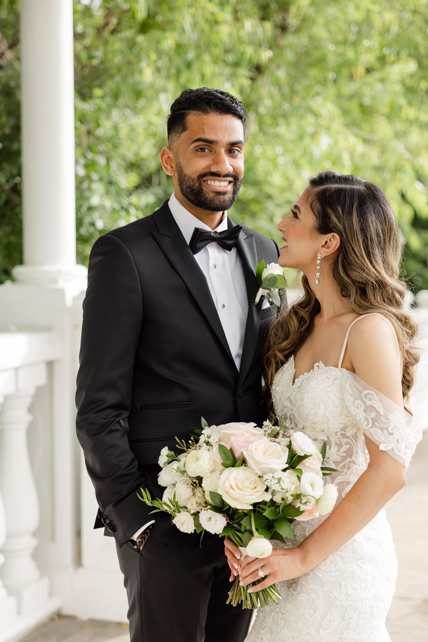 Wedding couple poses beneath white column with gorgeous bouquet of ivory roses.