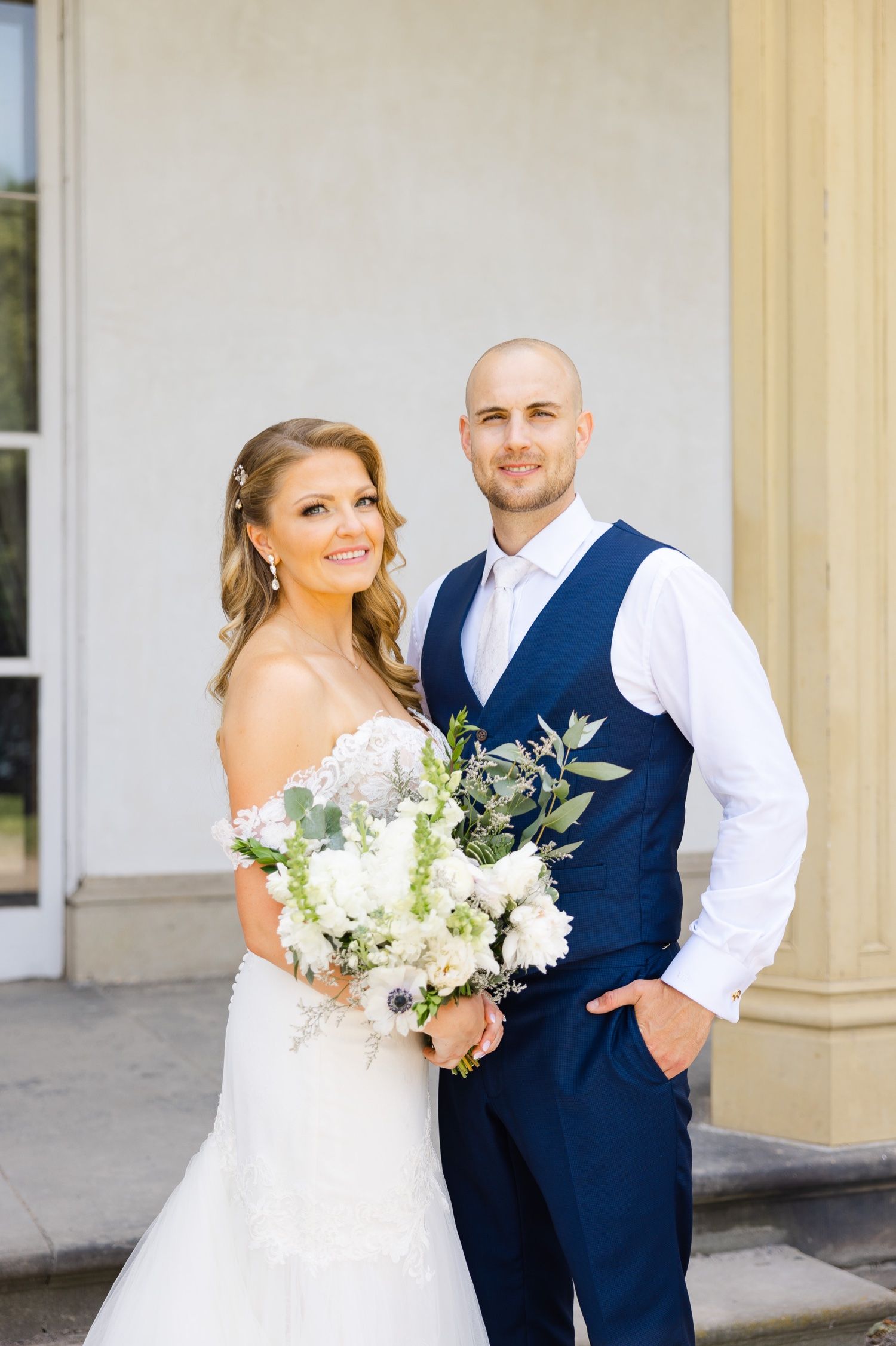 A wedding couple poses together on a porch, with the bride holding a white bouquet and groom wearing a navy vest.