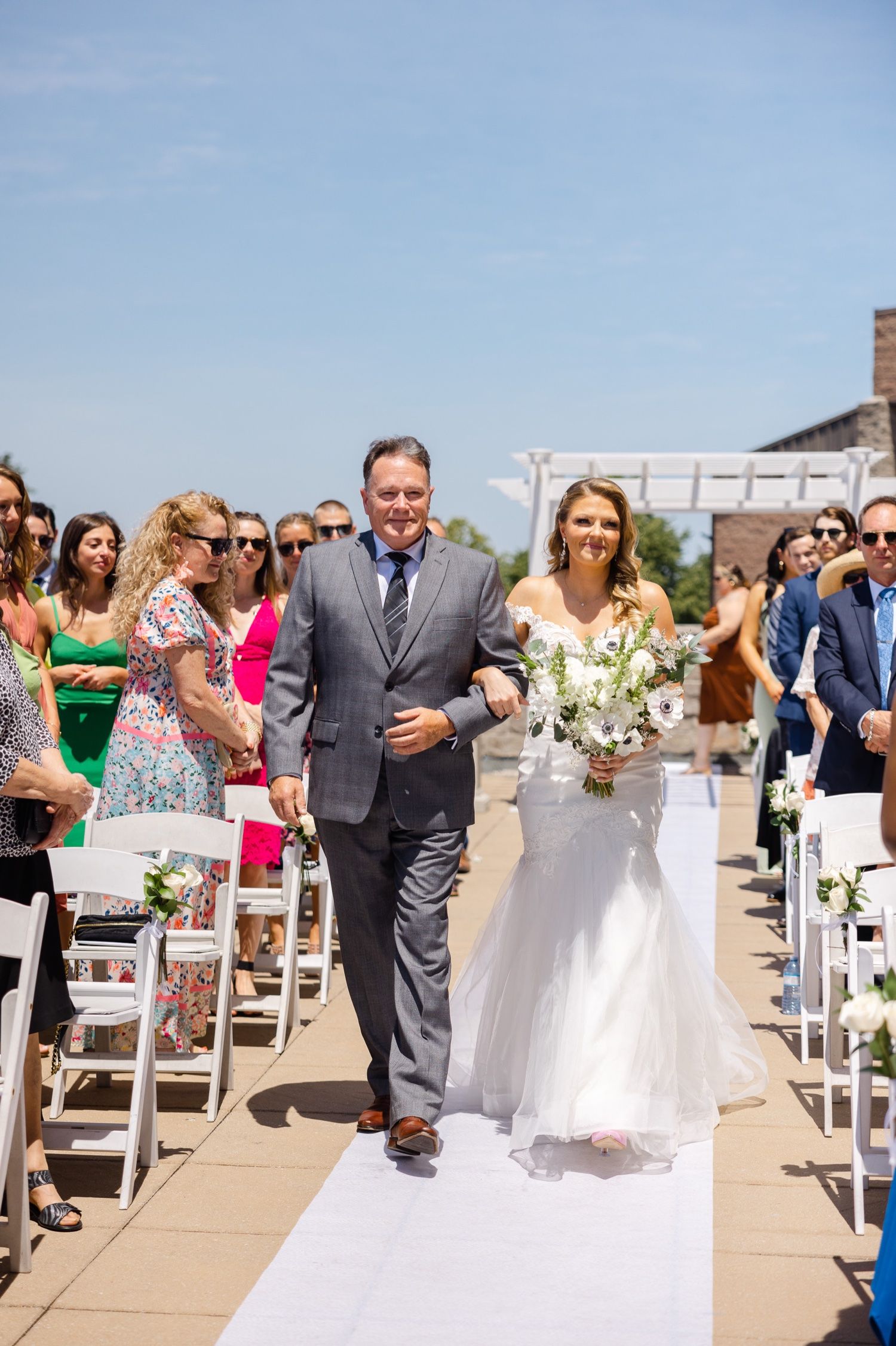 Wedding processional on white aisle runner with guests watching from white chairs.