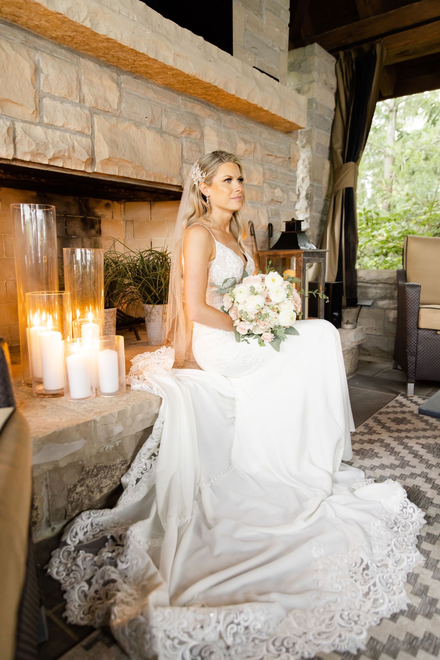 Elegant bride in lace dress sitting by candlelit stone fireplace.