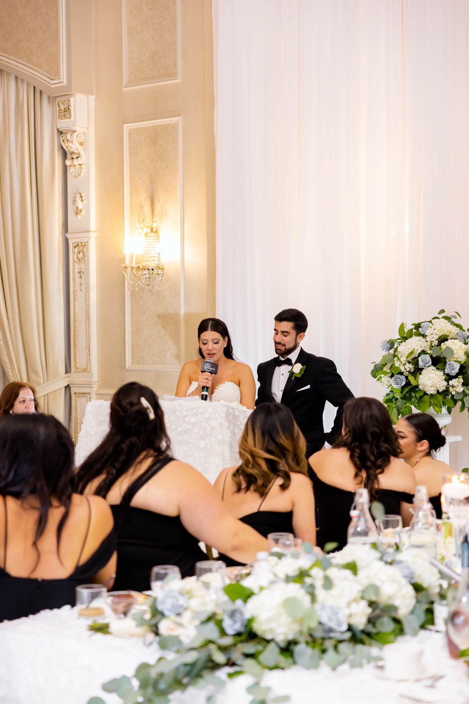 Newlyweds address their guests from decorative sweetheart table as bridesmaids listen.