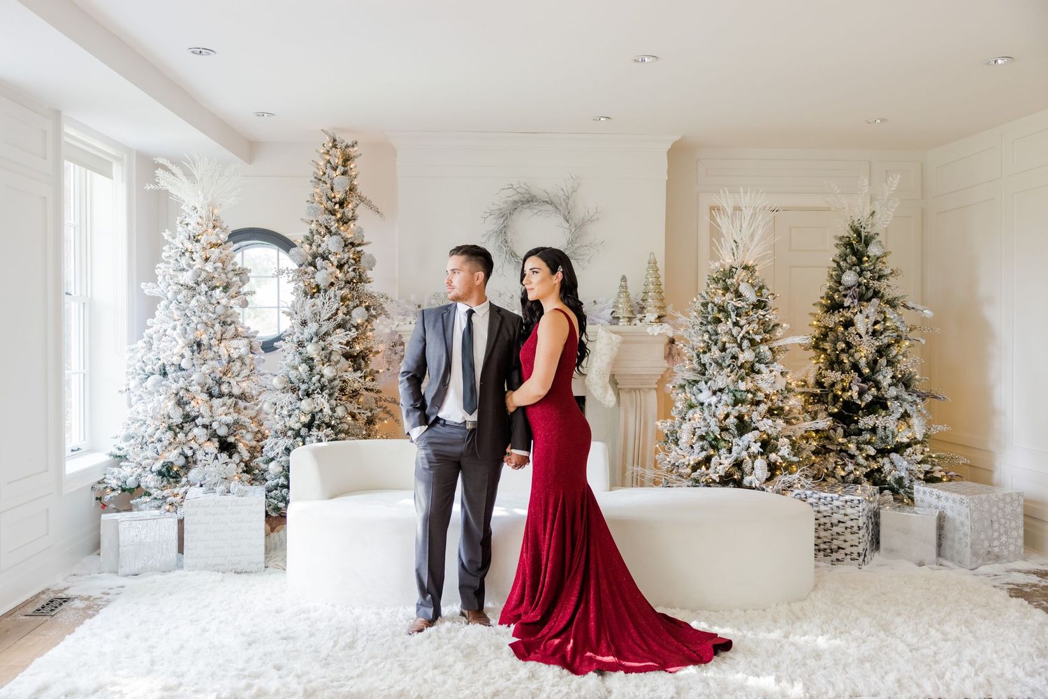 Elegant couple in formal attire pose in winter wonderland setting with decorated Christmas trees.