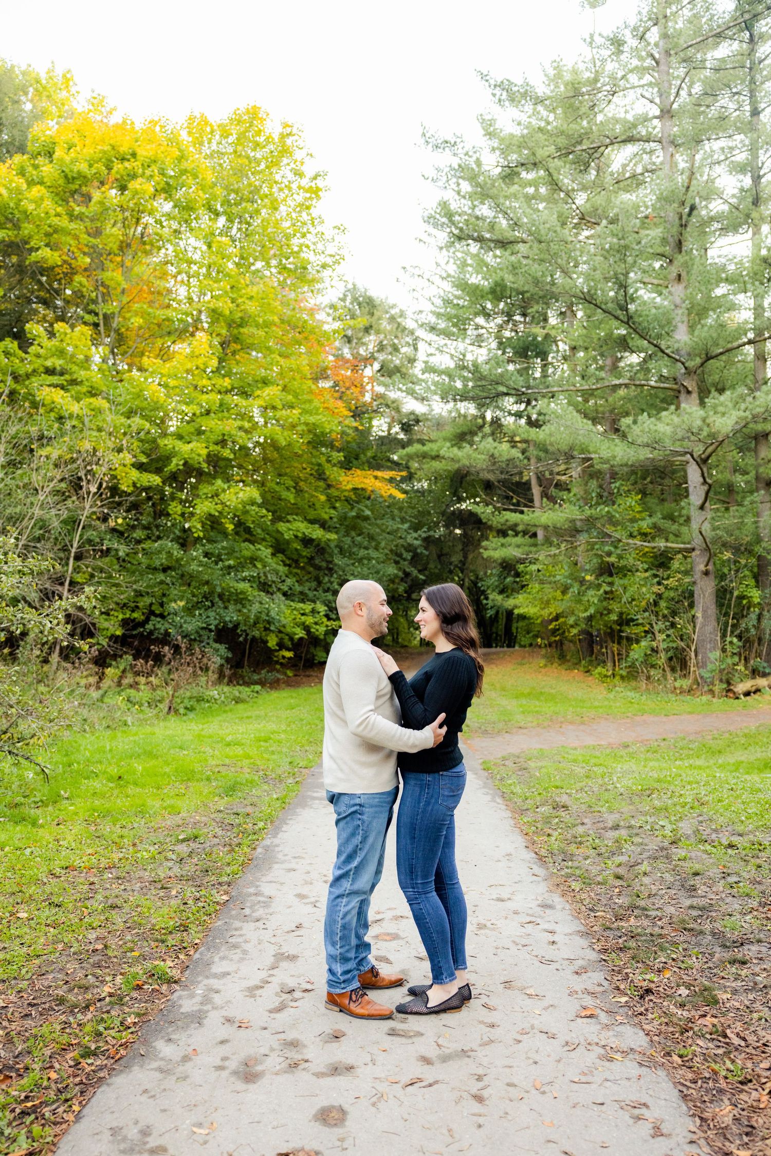 A couple embraces on a path surrounded by colorful fall foliage and evergreen trees.