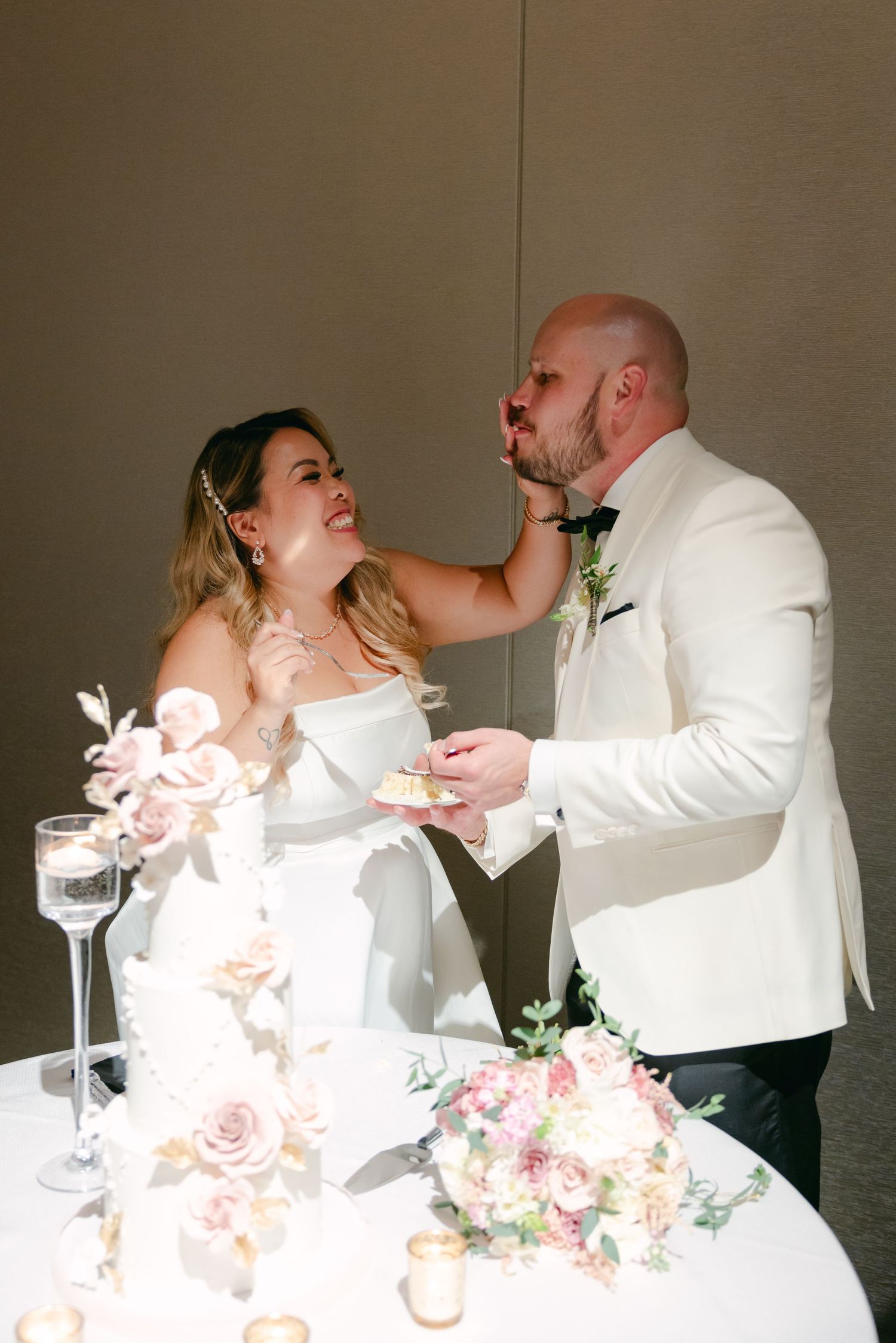 Newlyweds share a playful moment while cutting their wedding cake adorned with pink roses and floral decorations.