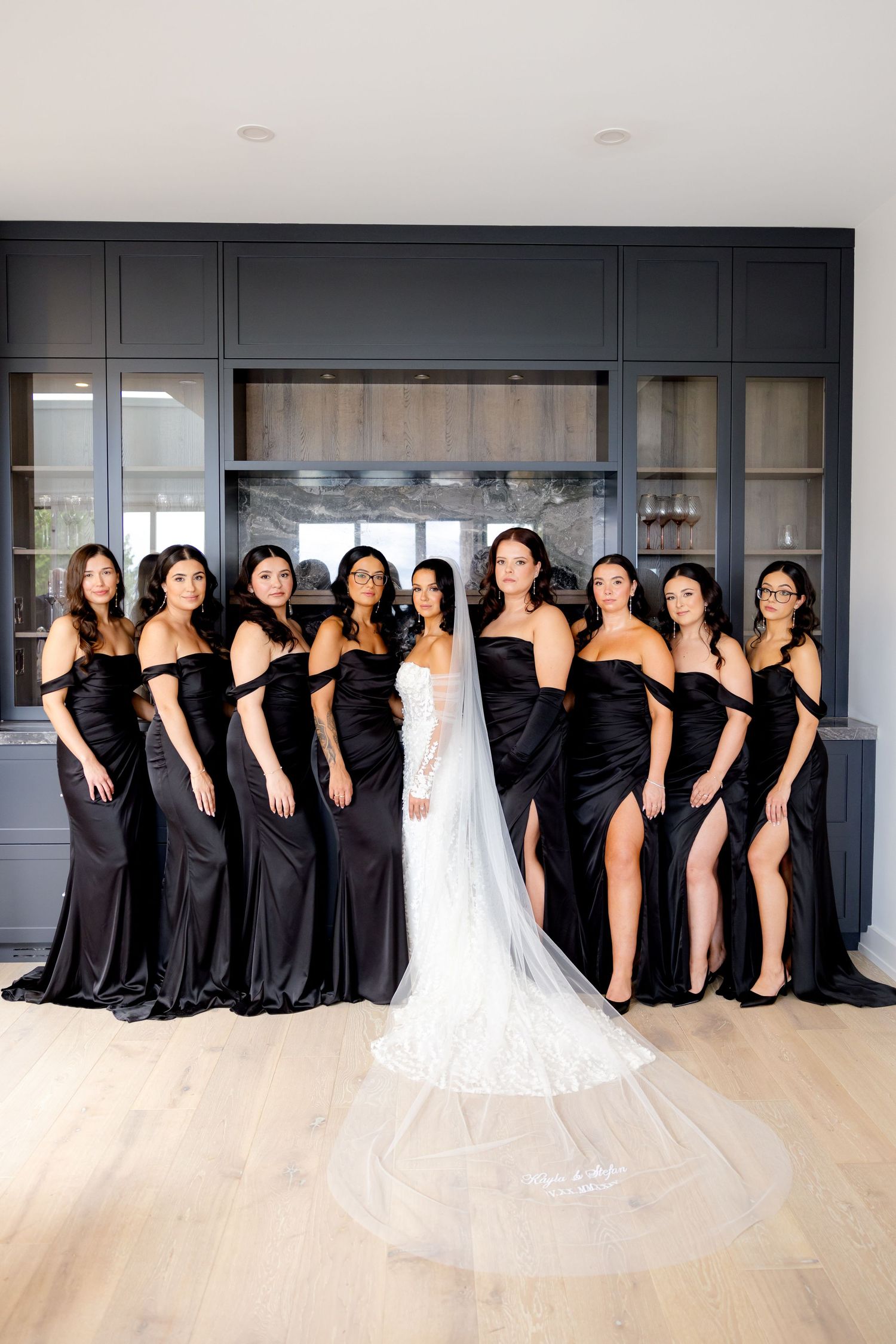Bridesmaids wearing elegant black gowns pose with bride in white wedding dress against modern grey built-in cabinet.