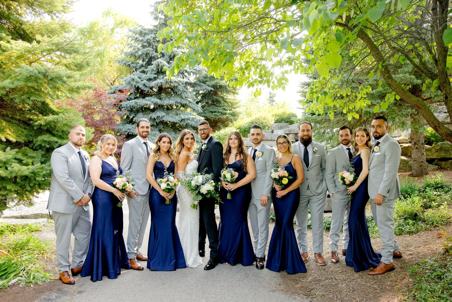 Wedding party in coordinated navy and gray attire poses on garden path with evergreen trees.