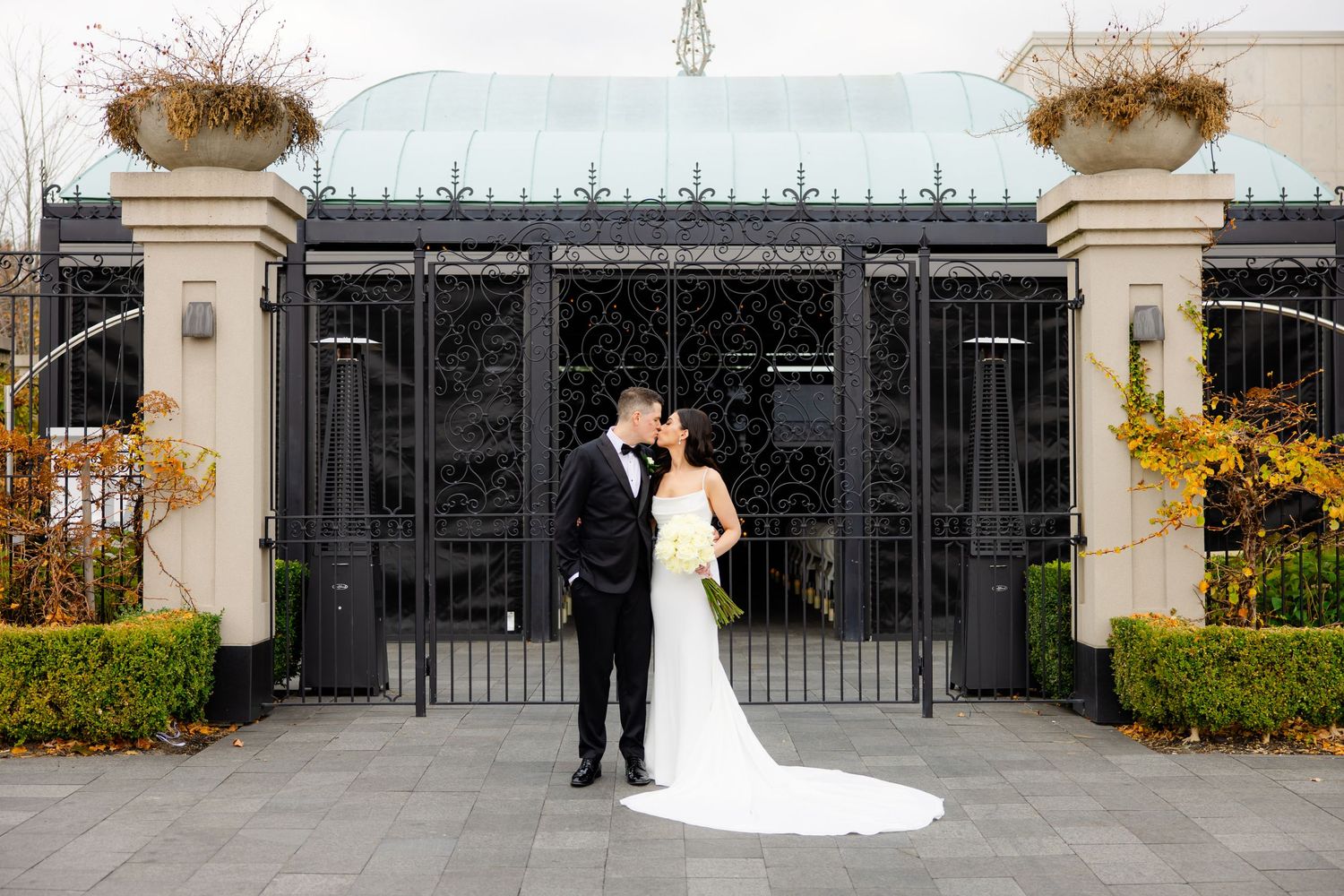 Wedding couple poses in front of grand black gates with decorative stone pillars and autumn foliage.
