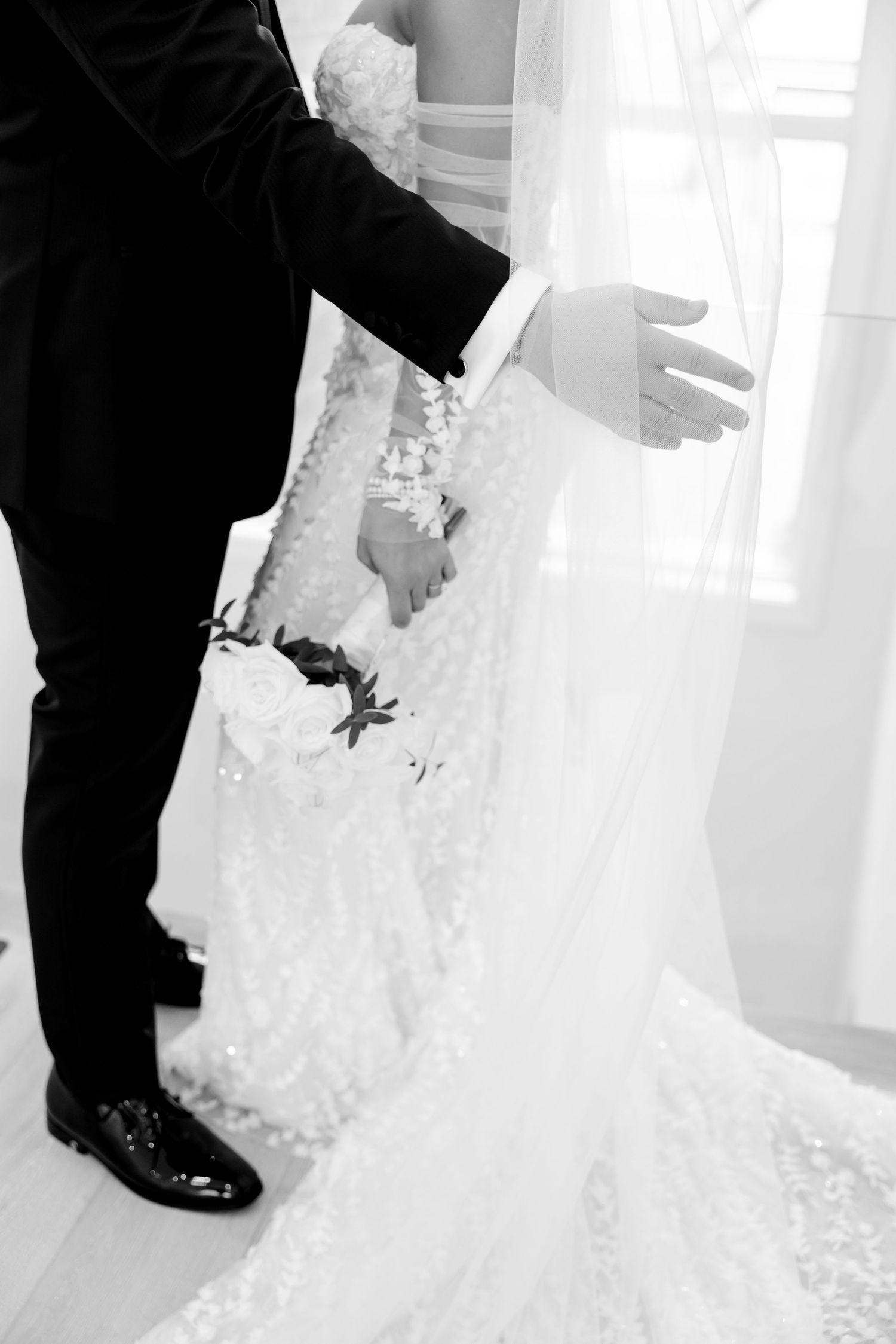 Close-up detail shot of wedding dress train and groom's suit in an elegant white setting.