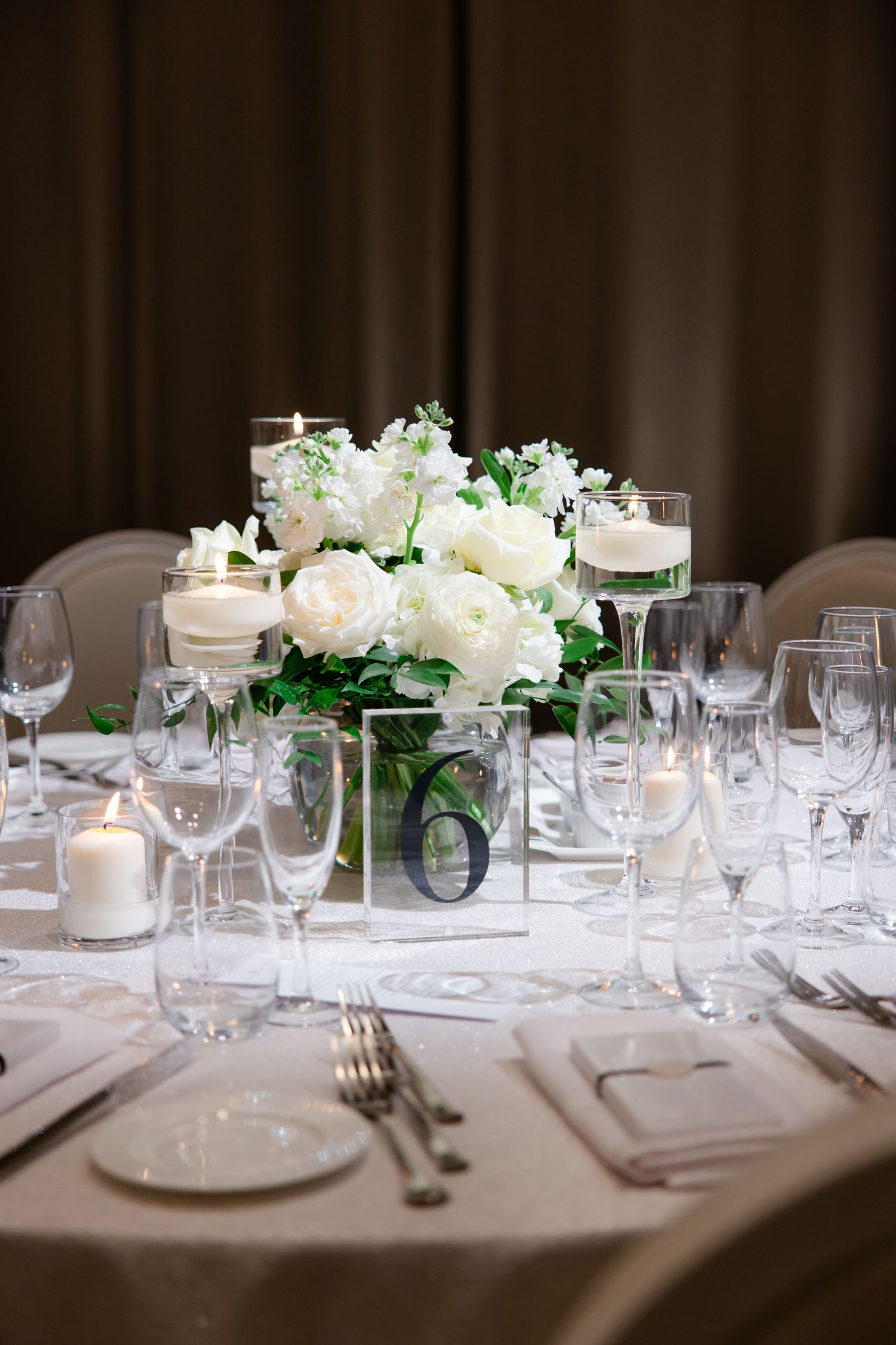 Formal table setting with white roses in glass vase centerpiece and table number six.