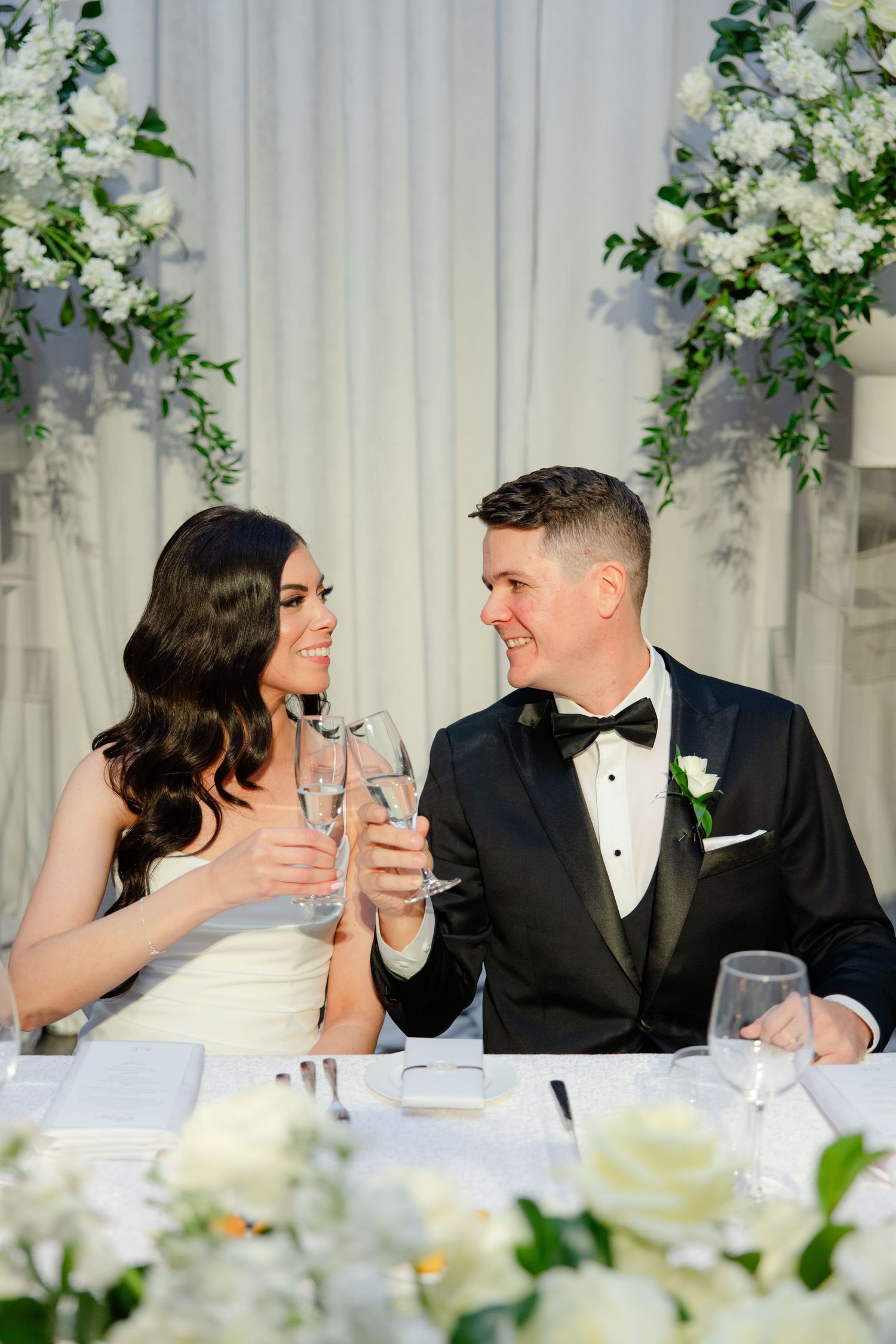 Couple toasts with champagne at their wedding reception head table decorated with white floral arrangements.