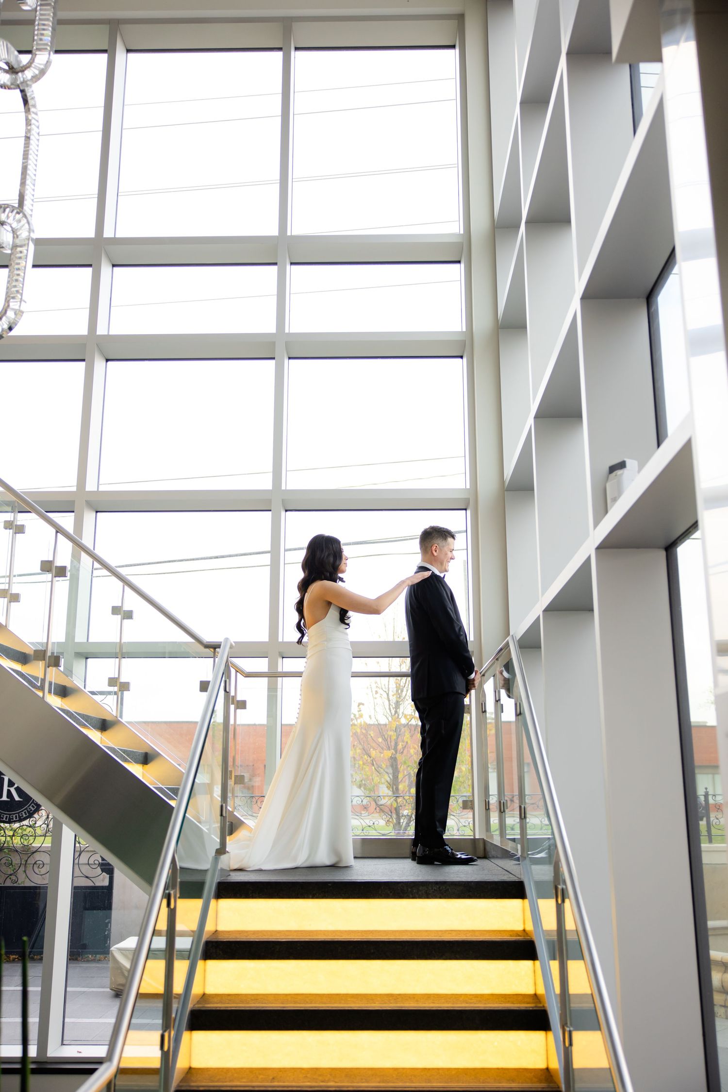 Couple in wedding attire stands back-to-back on modern yellow staircase with industrial decor.
