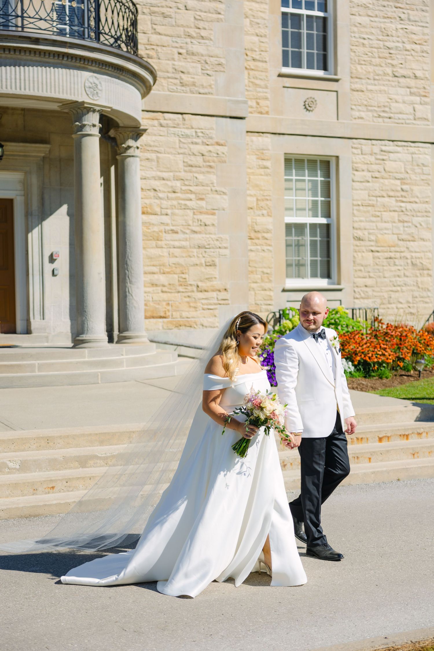 A couple in wedding attire poses outside an elegant stone mansion with columned entrance.