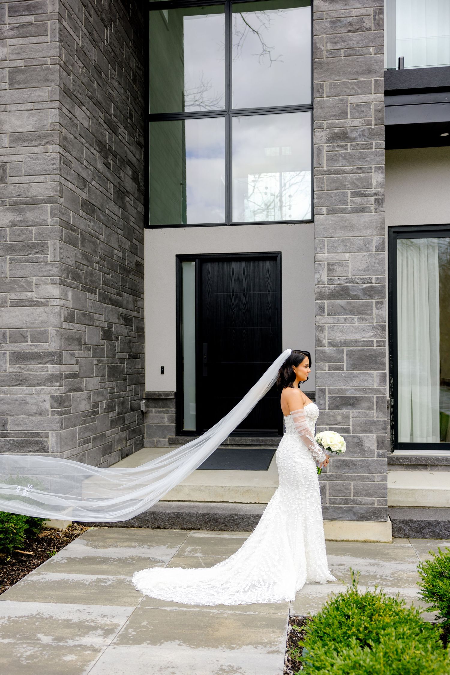 A bride in a fitted lace wedding dress with cathedral veil poses outside a modern building.