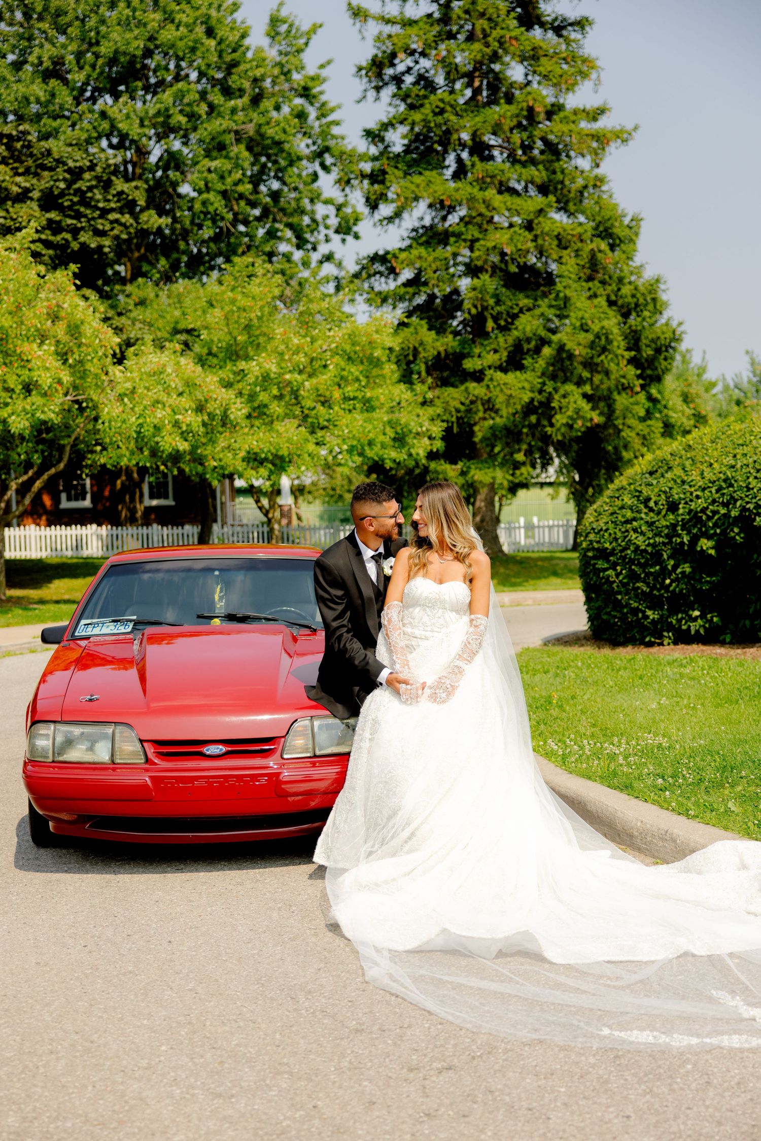 Newlyweds pose for romantic wedding photos next to a red vintage Mustang car in a sunny park setting.