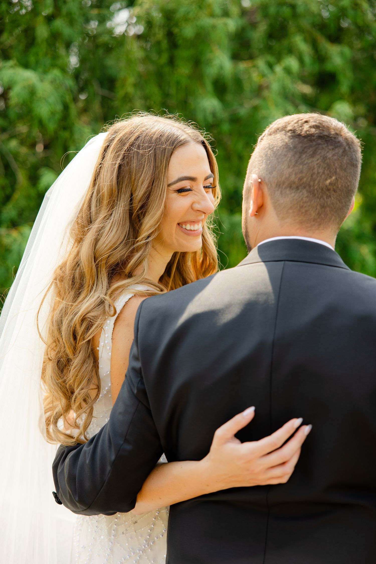 A joyful bride in a white dress smiles while embracing her groom outdoors against lush greenery.