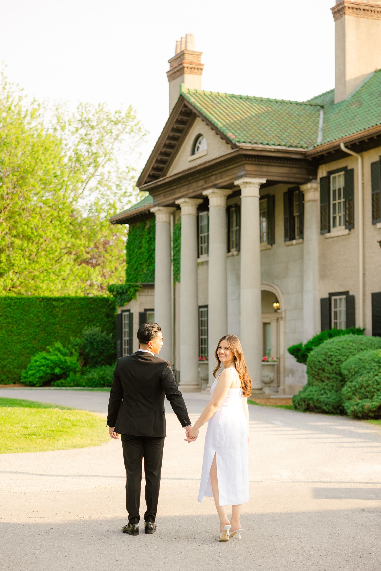 Couple walking hand-in-hand toward stately mansion with green roof and white columns.