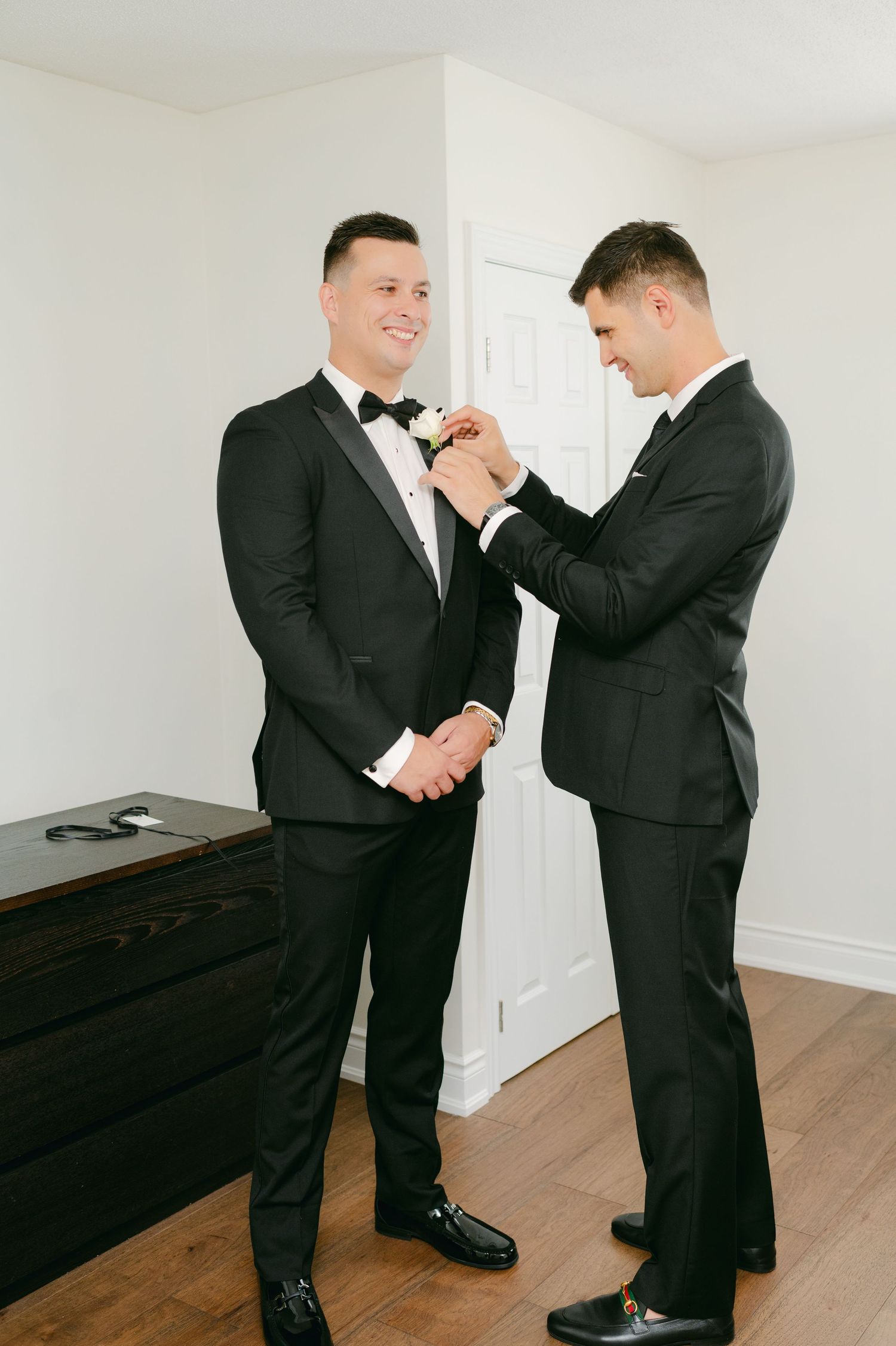 Two men in tuxedos during wedding preparations as one assists the other with his bowtie.