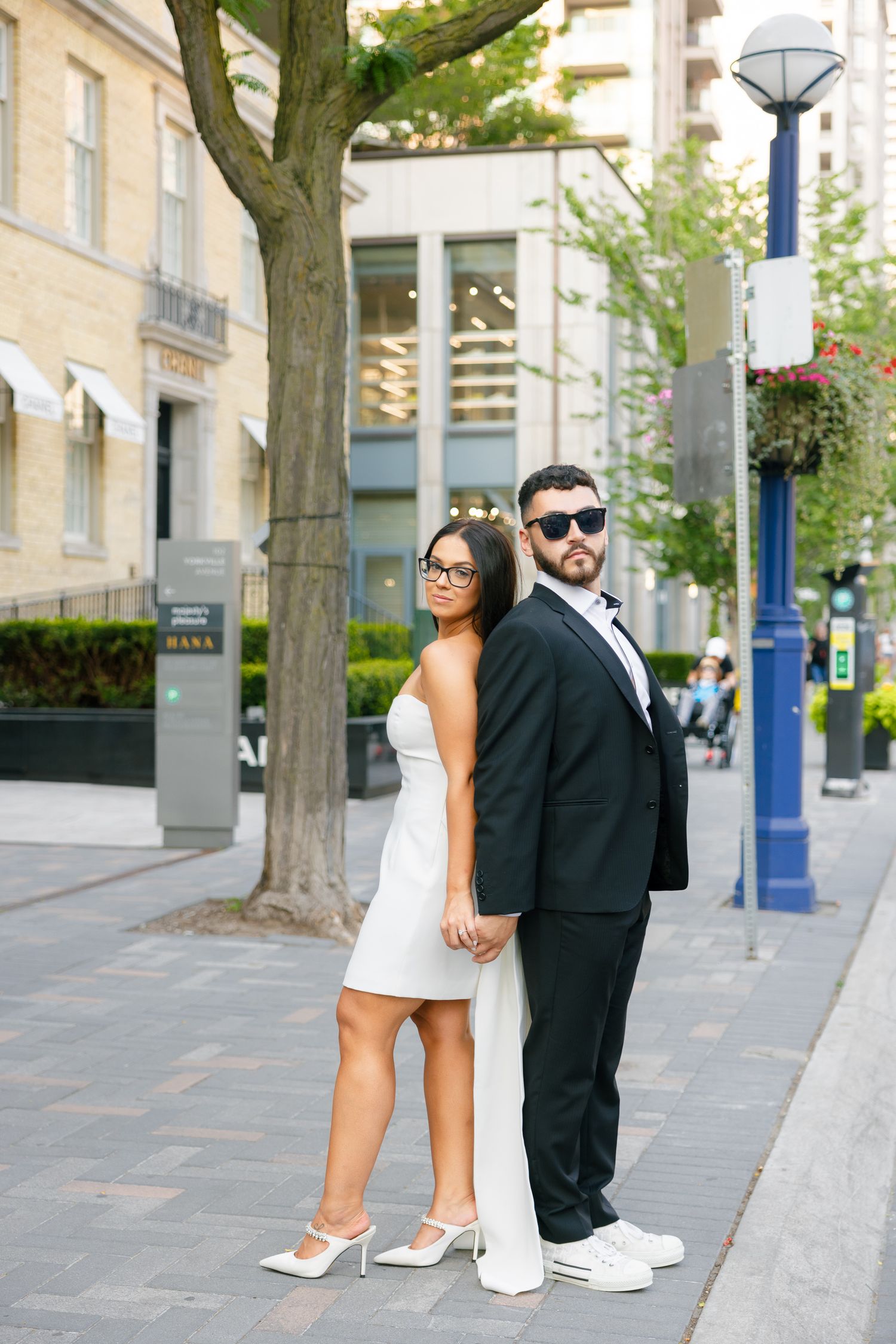 A couple poses on a city sidewalk in contrasting formal attire, with white sneakers and a modern urban backdrop.