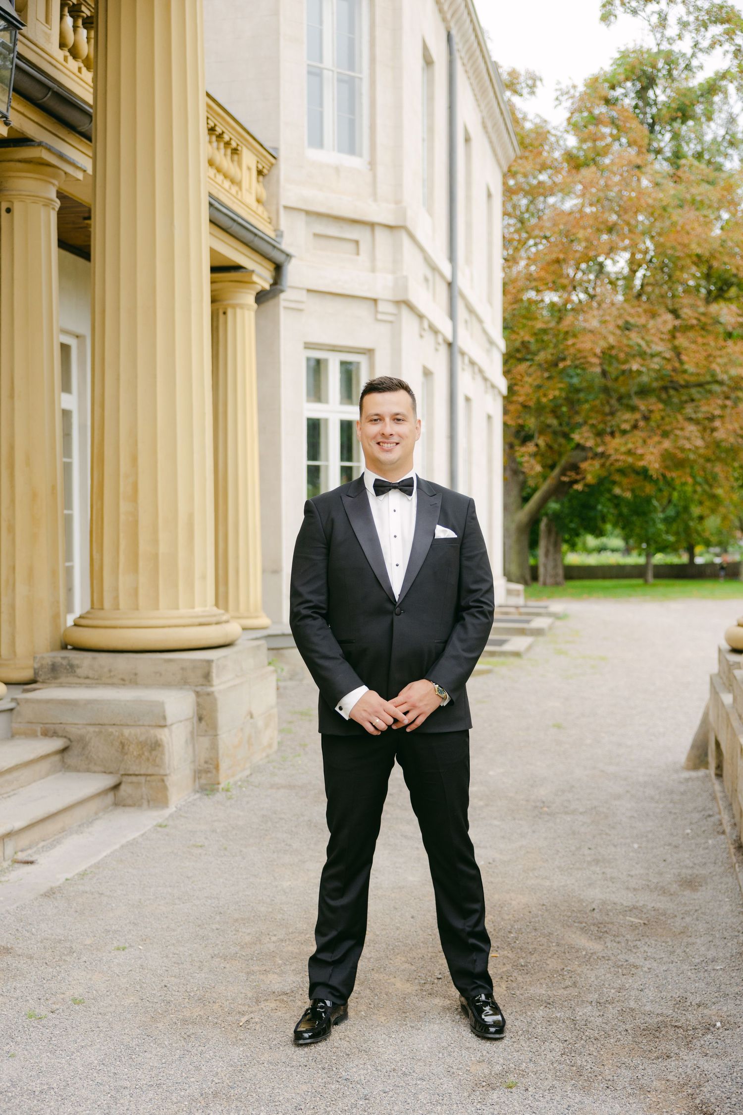 Groom in classic black tuxedo stands confidently in front of grand historic building.