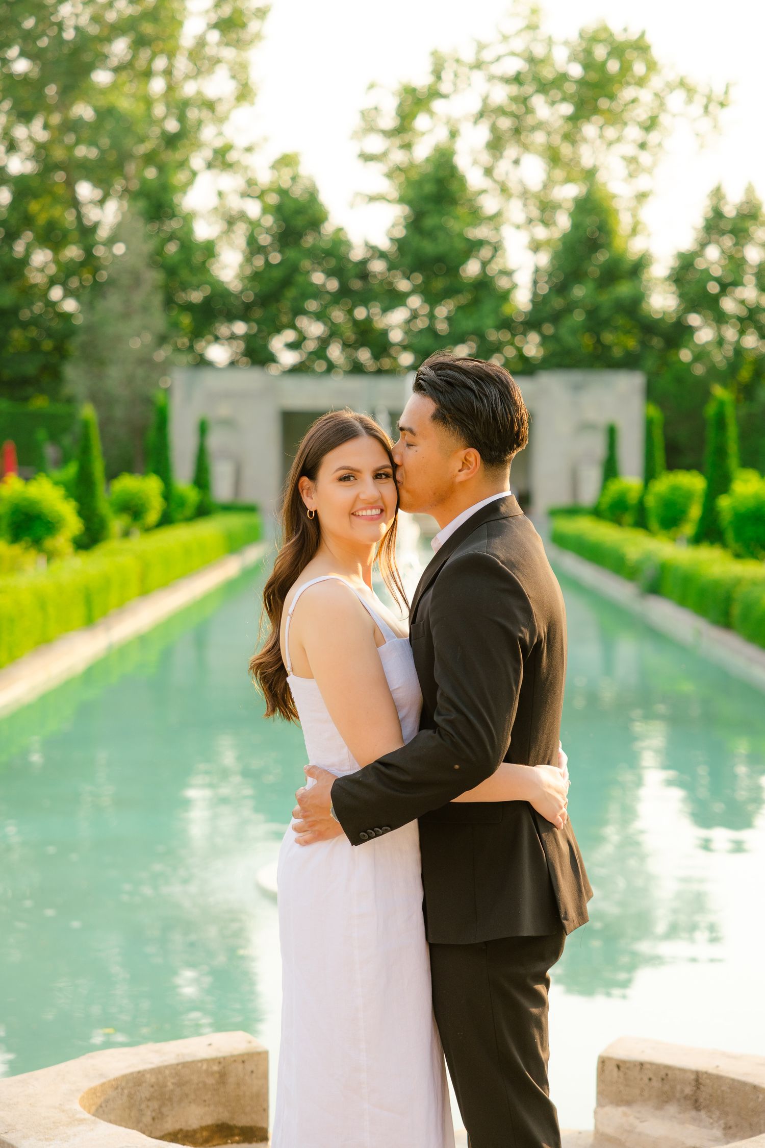 A couple sharing an intimate embrace in a sunlit garden with a reflecting pool.