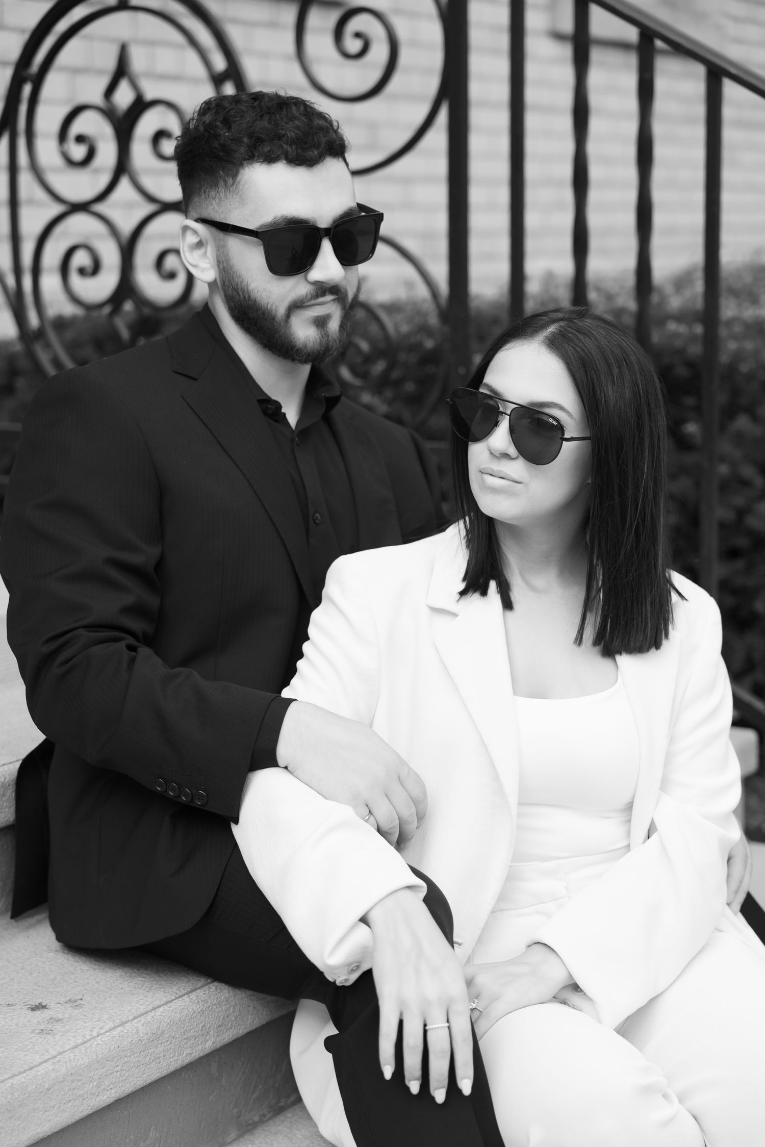 Black and white portrait of couple in designer outfits against ornate iron railing.
