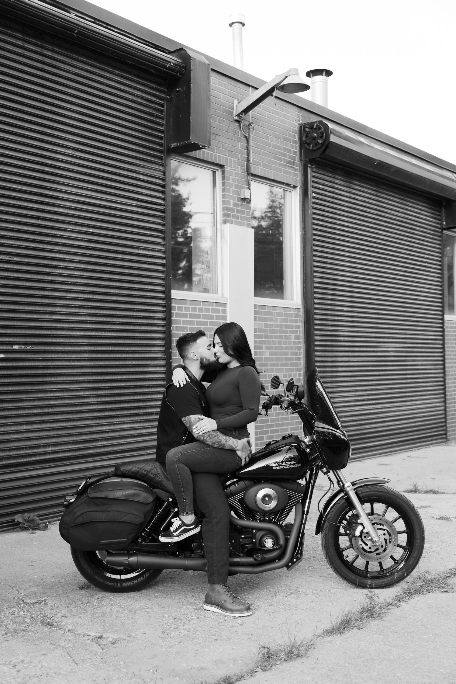 A couple shares a romantic moment on their motorcycle outside an industrial building.