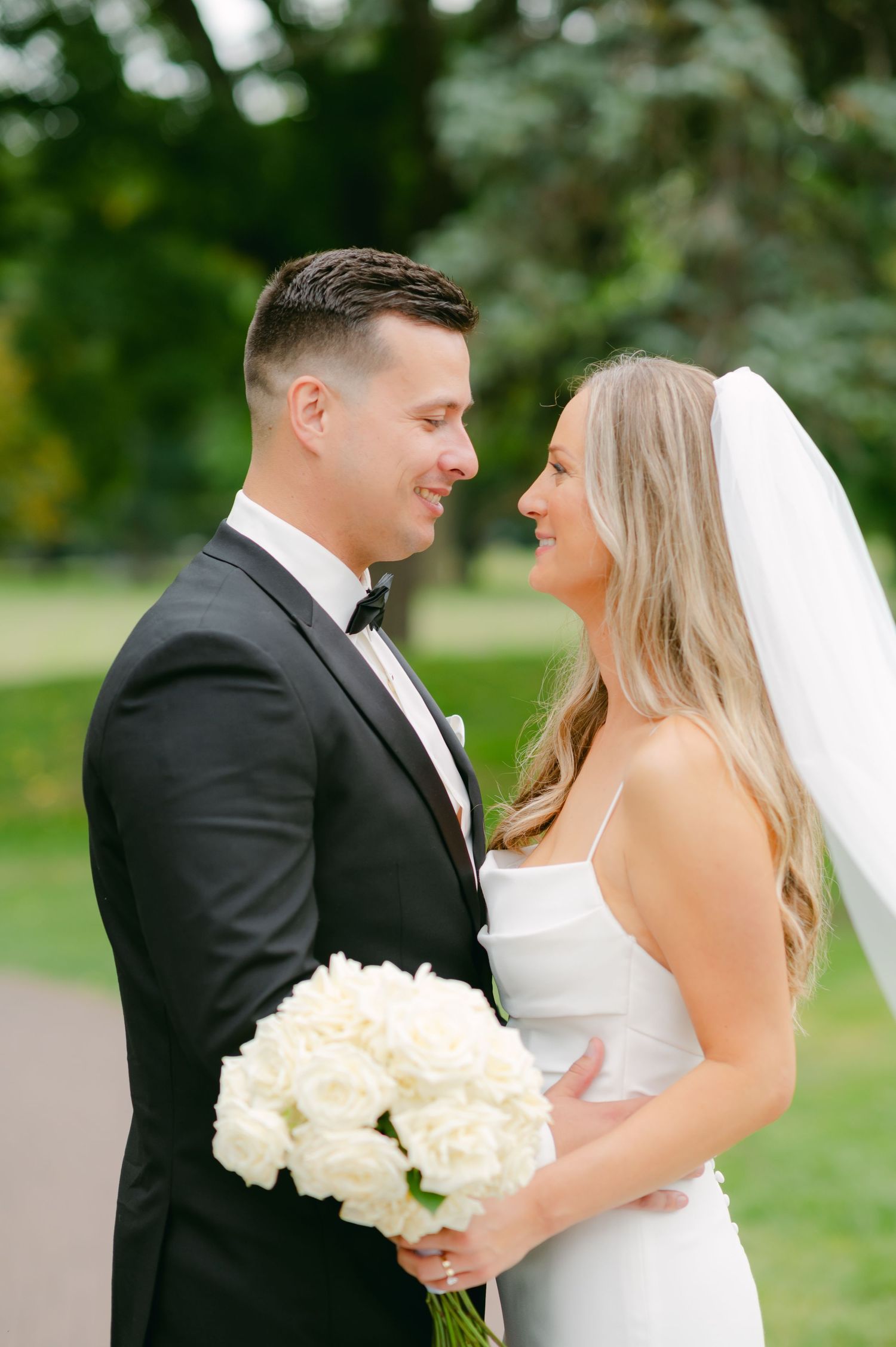 Wedding couple sharing joyful moment outdoors while holding elegant white rose bouquet.