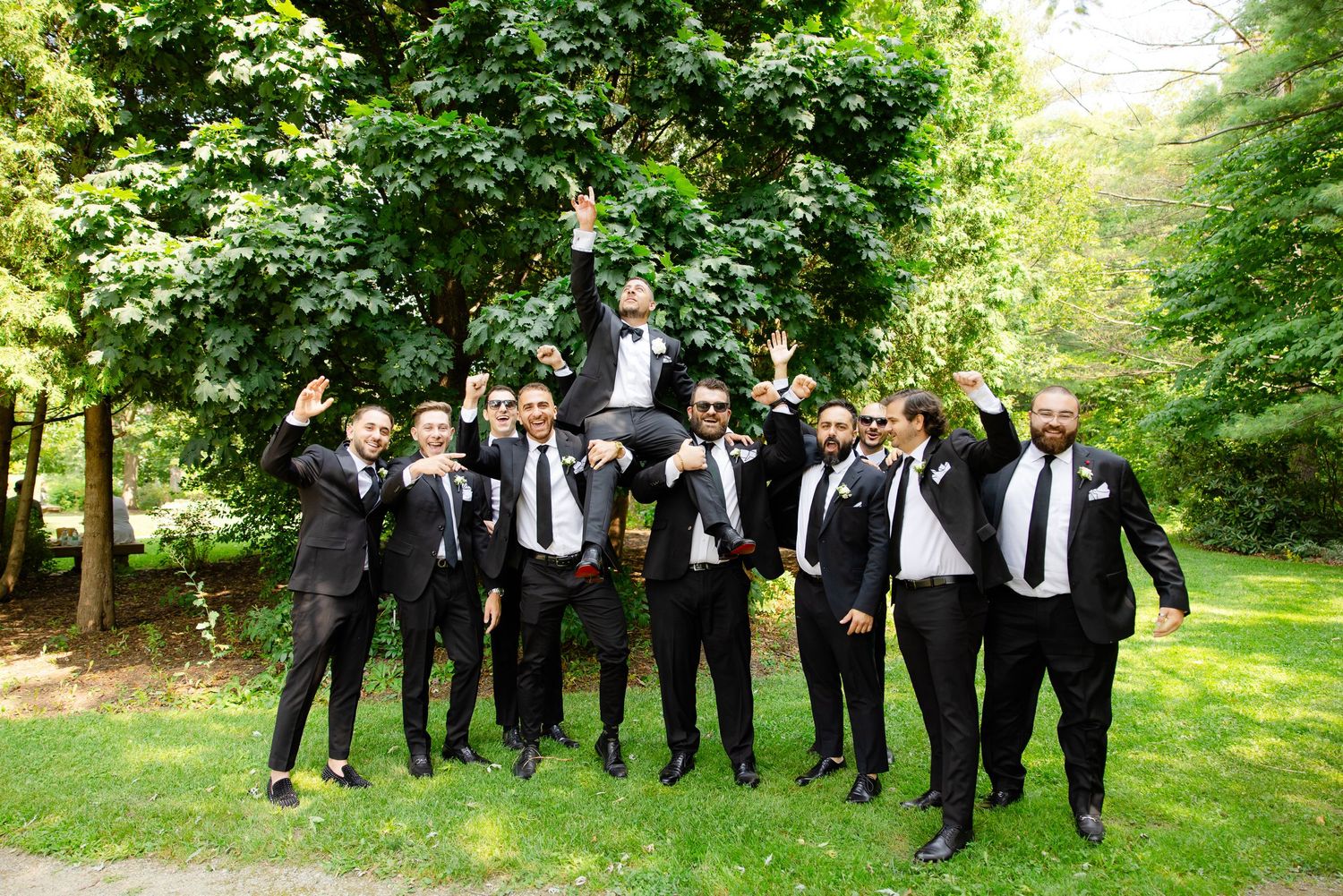 Groomsmen in black suits celebrating joyfully outdoors in a forest setting.