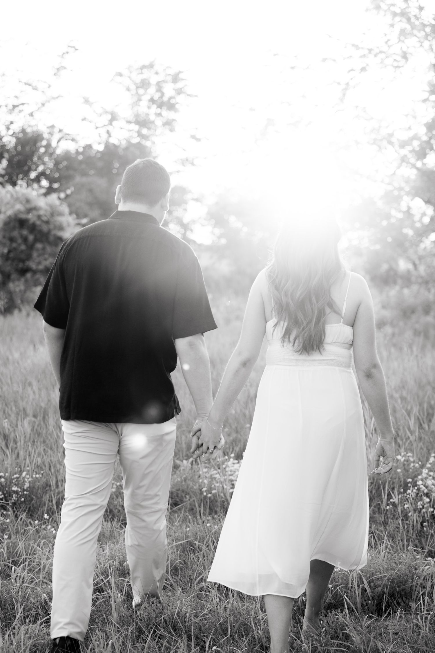 Black and white silhouette of couple walking through meadow at sunset holding hands.