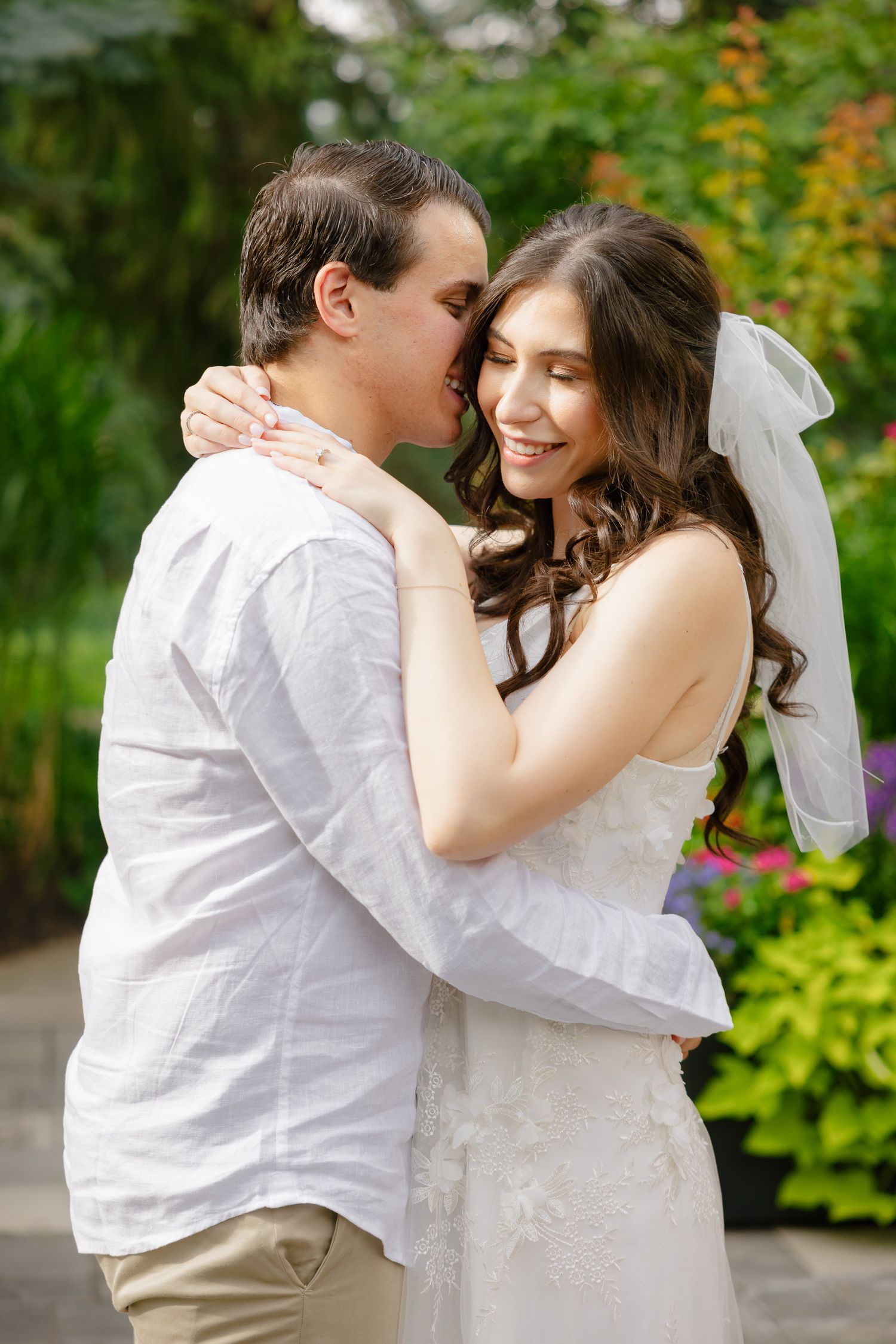 A tender moment between newlyweds embracing in a garden with evergreens and flowers in the background.